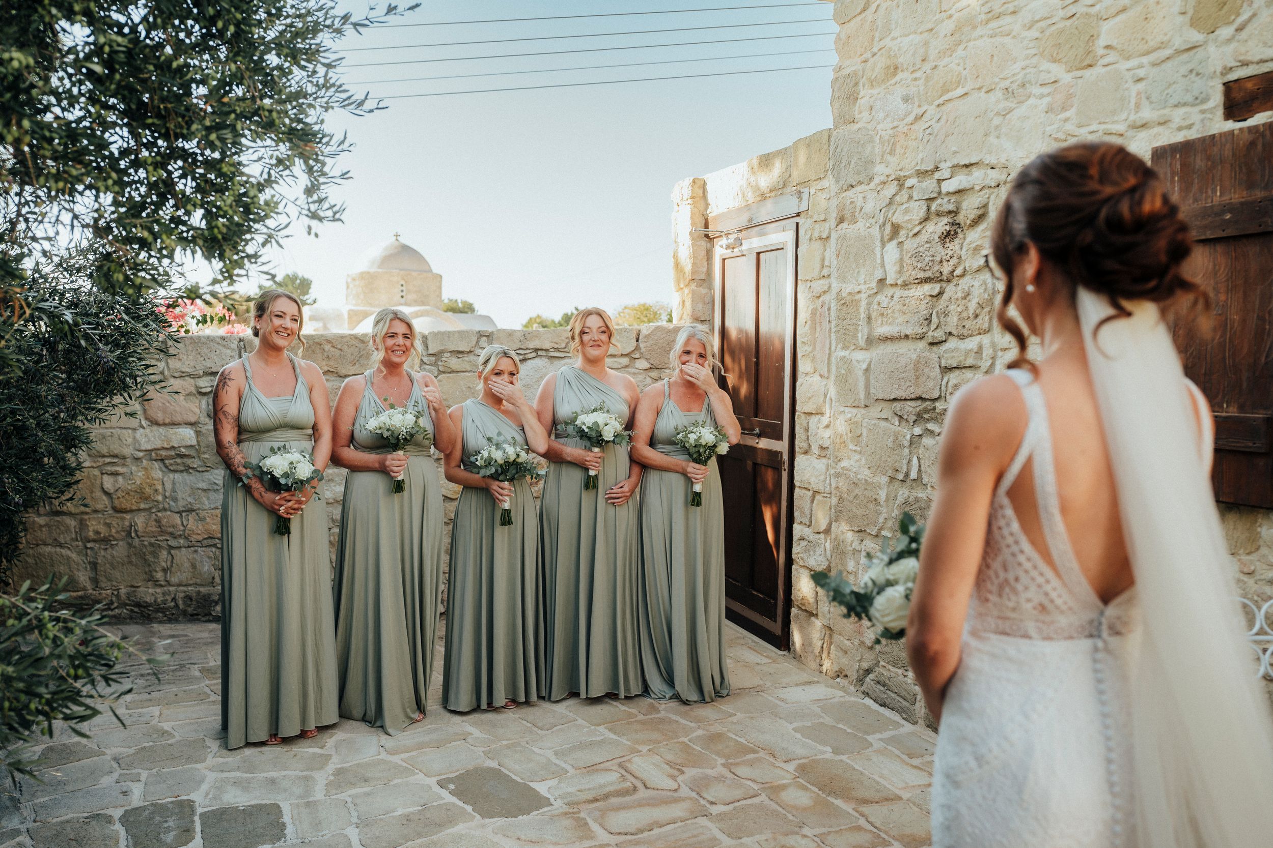 A wedding party in sage green dresses poses together against a rustic stone wall at an outdoor venue.