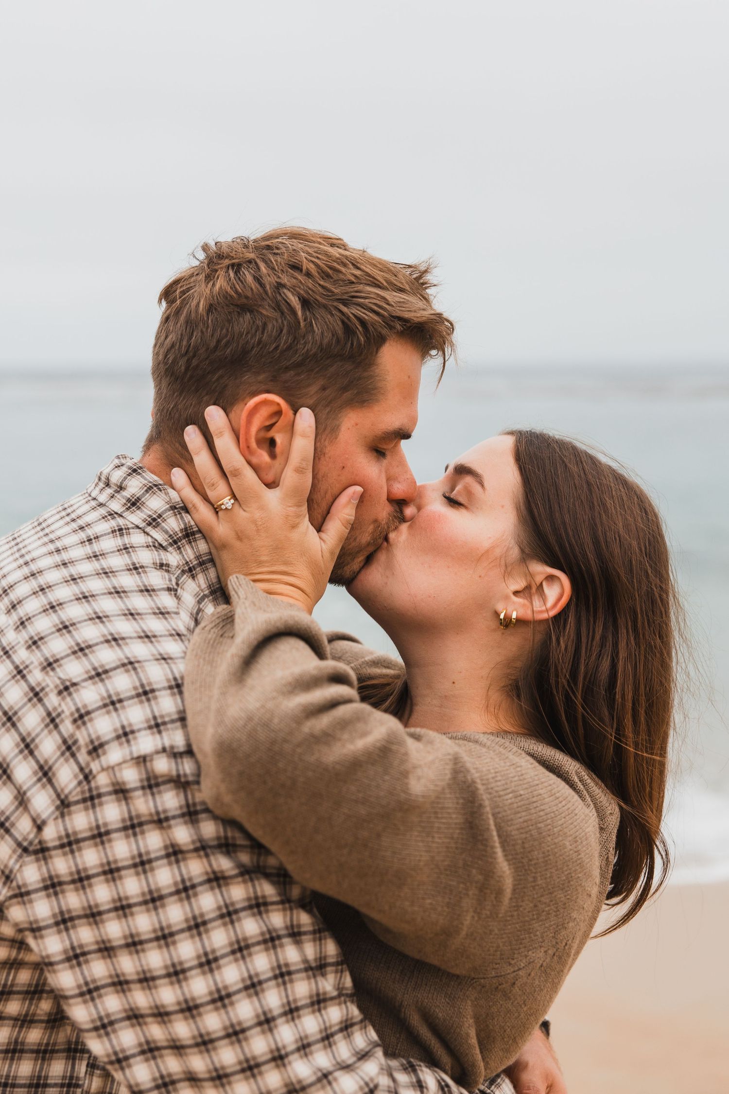 A romantic couple sharing an intimate kiss on a cloudy beach while wearing casual fall attire.