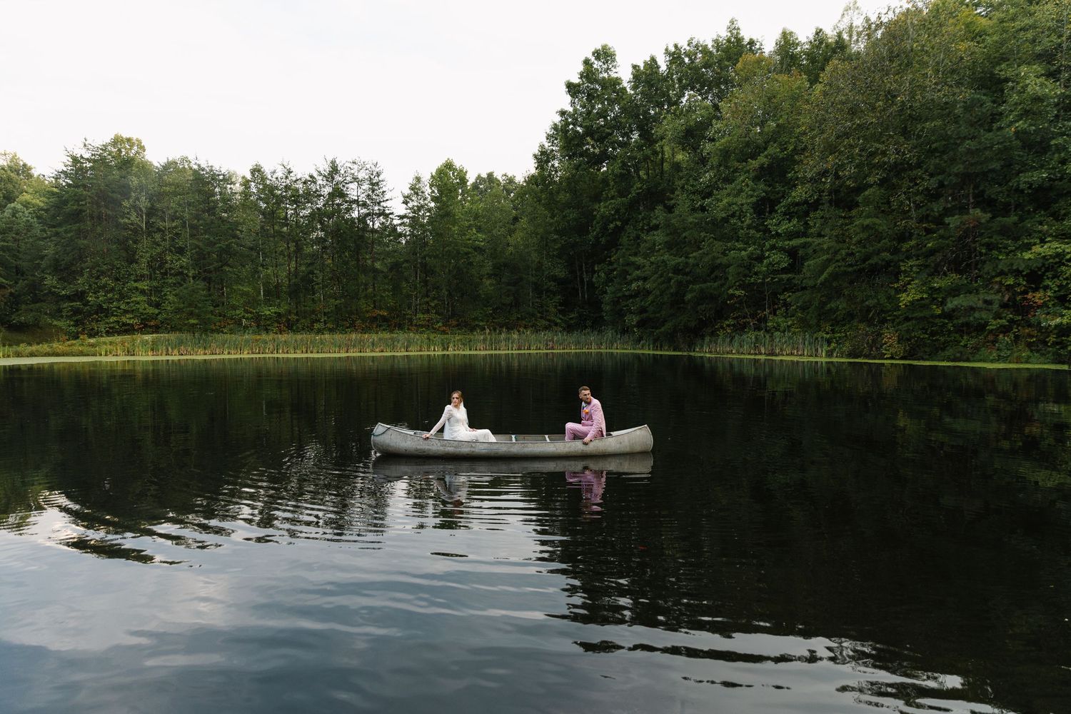 Two people in wedding attire paddle across a tranquil lake in a silver canoe.