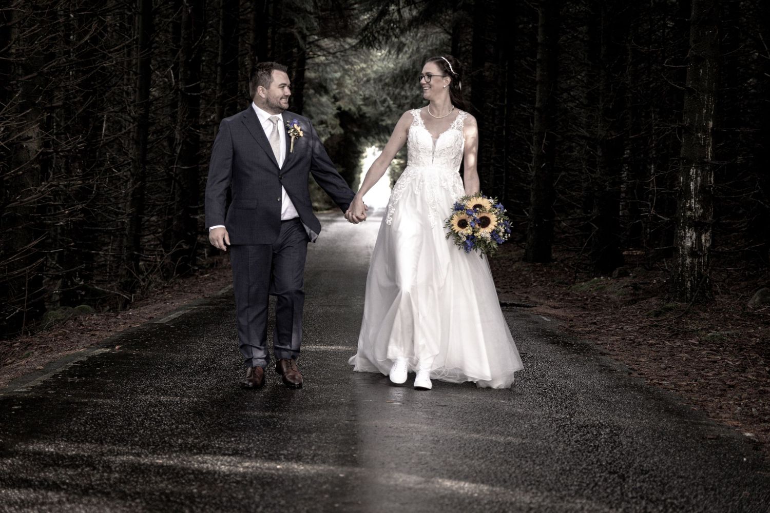 Bride and groom walking hand in hand down a dark forest road on their wedding day.