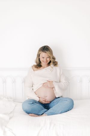 Expectant mother in a cozy white sweater and jeans sits cross-legged on white couch looking down at her belly.