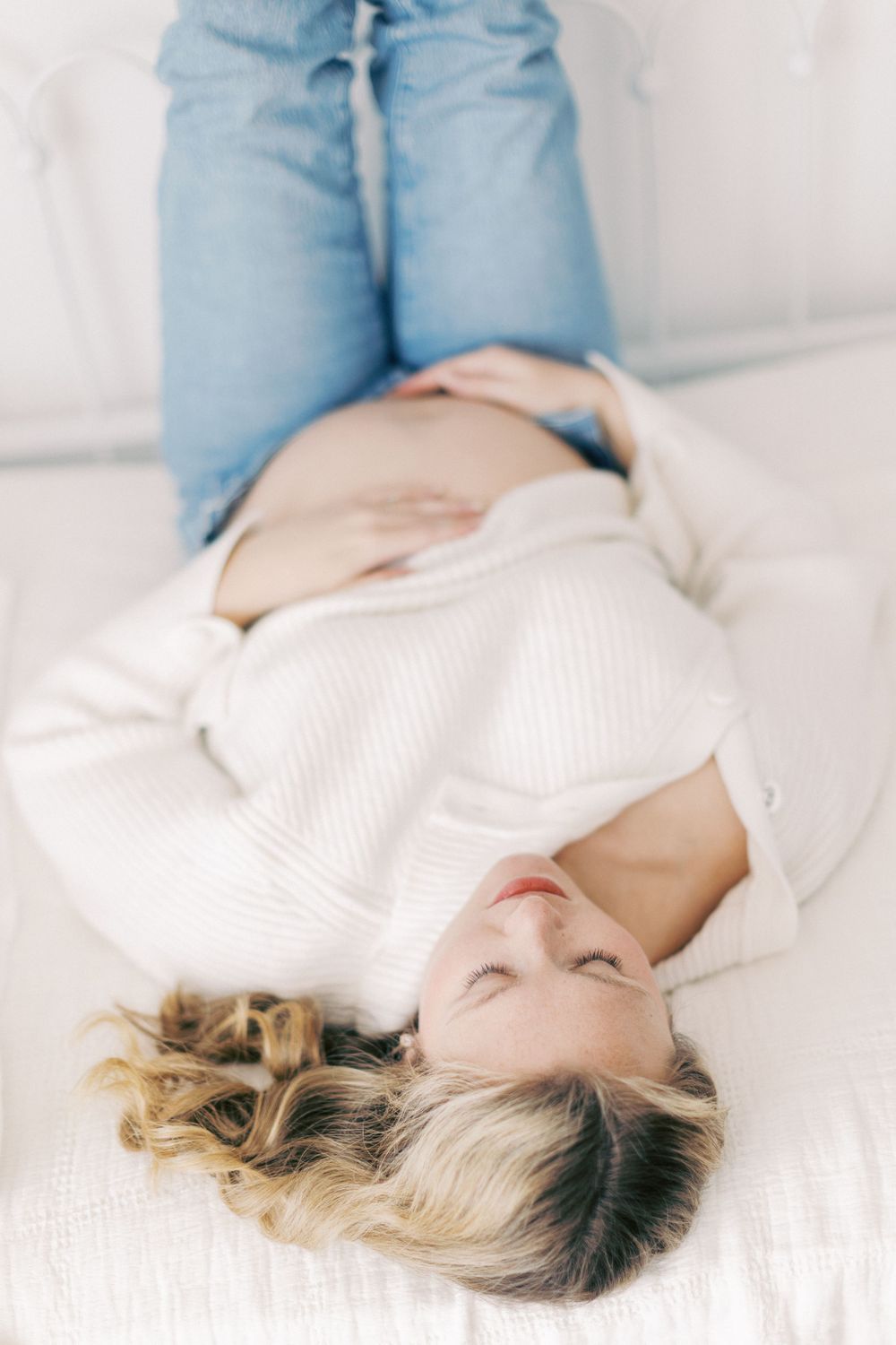 Someone with blonde hair lying down on white bedding wearing a cozy white sweater and light blue jeans.