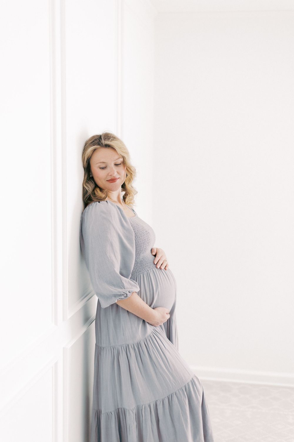 A person in a flowing grey dress cradles their pregnant belly while standing against a white wall in soft lighting.