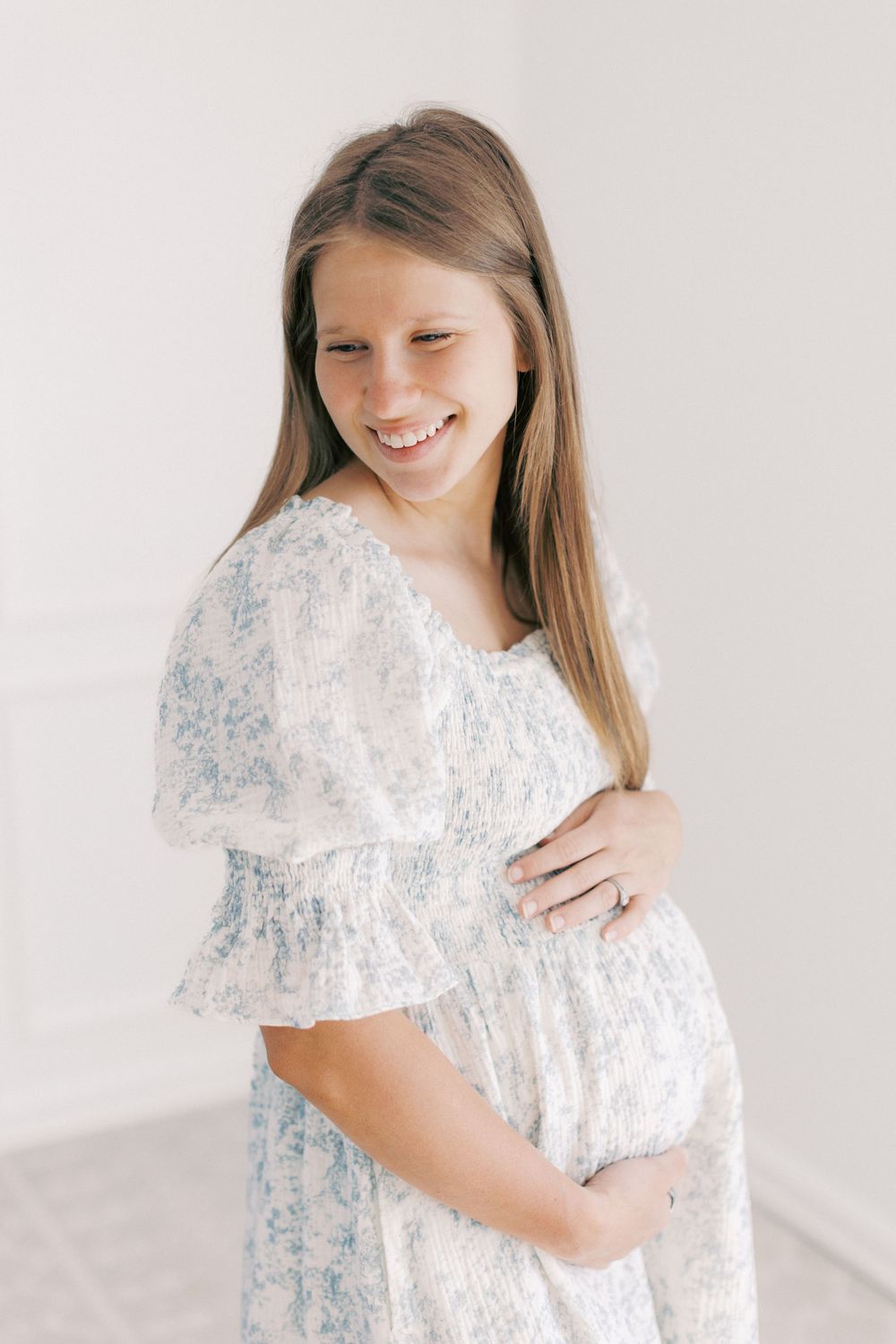 Smiling expectant mother in a white floral dress with ruffled sleeves poses against a light background.