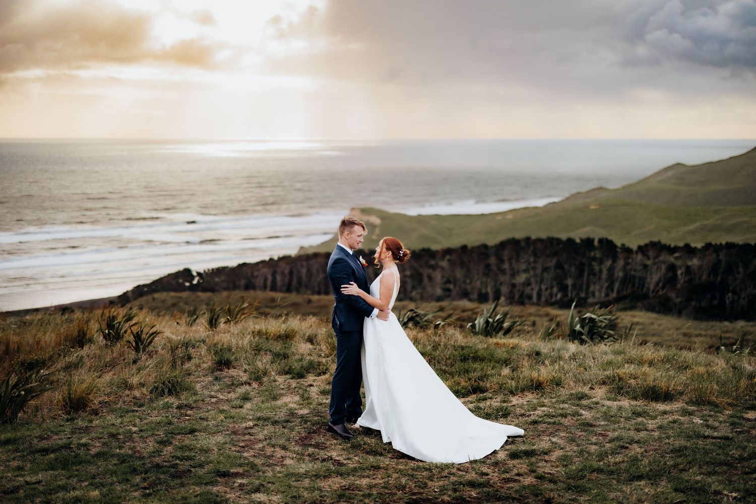 Dramatic coastal wedding portrait with couple standing on clifftop overlooking ocean at sunset.