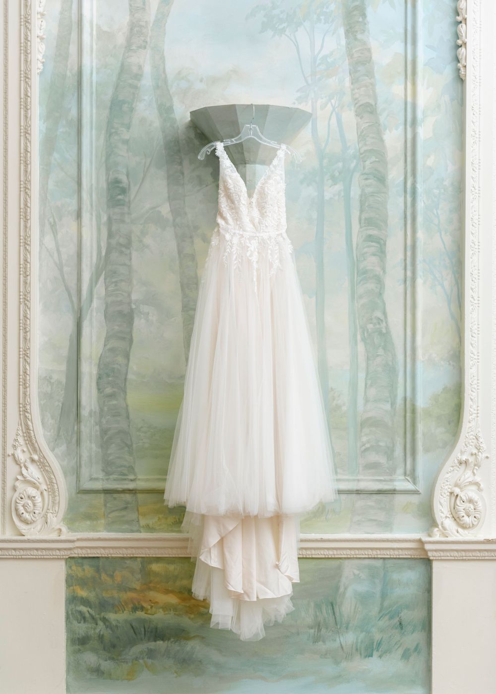 Elegant white tulle wedding gown with off-shoulder straps hangs displayed against a painted wall with birch tree motif.