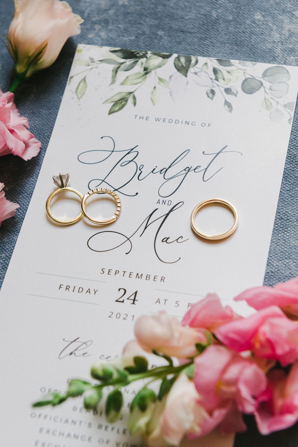 Wedding invitation with floral design and gold wedding rings alongside pink flowers on a painted background.
