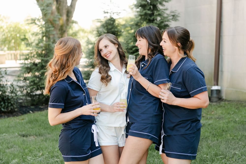 A bride and her bridesmaids in matching navy pajama sets laugh together while standing in a garden.