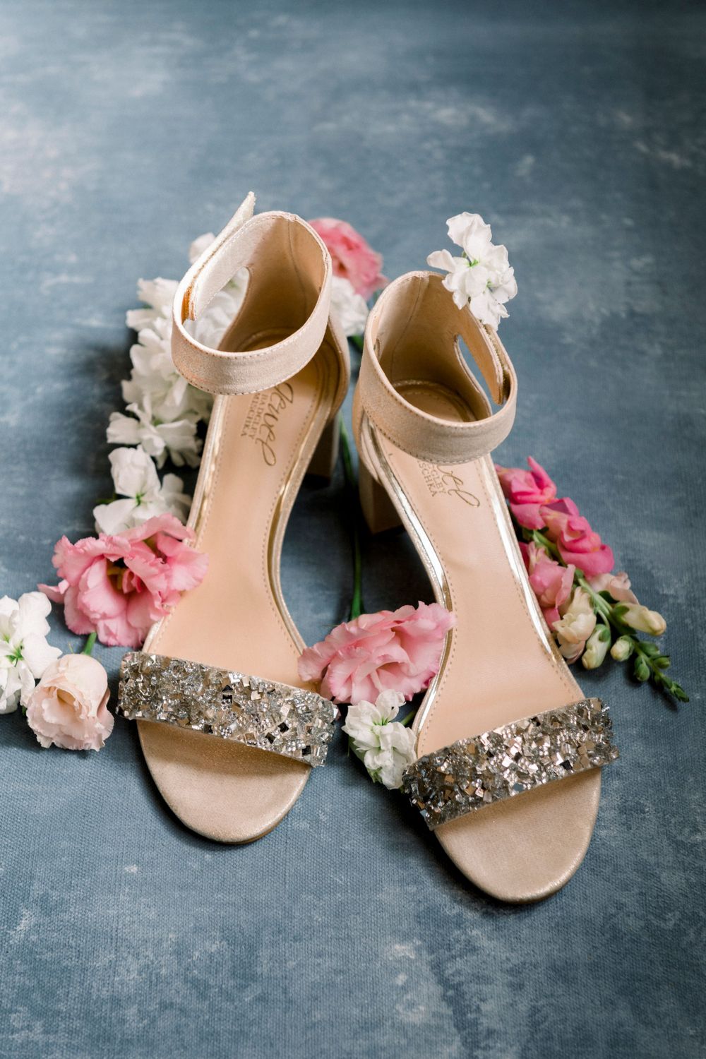 Elegant bridal shoes with sparkly straps displayed with pink and white flowers on a blue painted background.