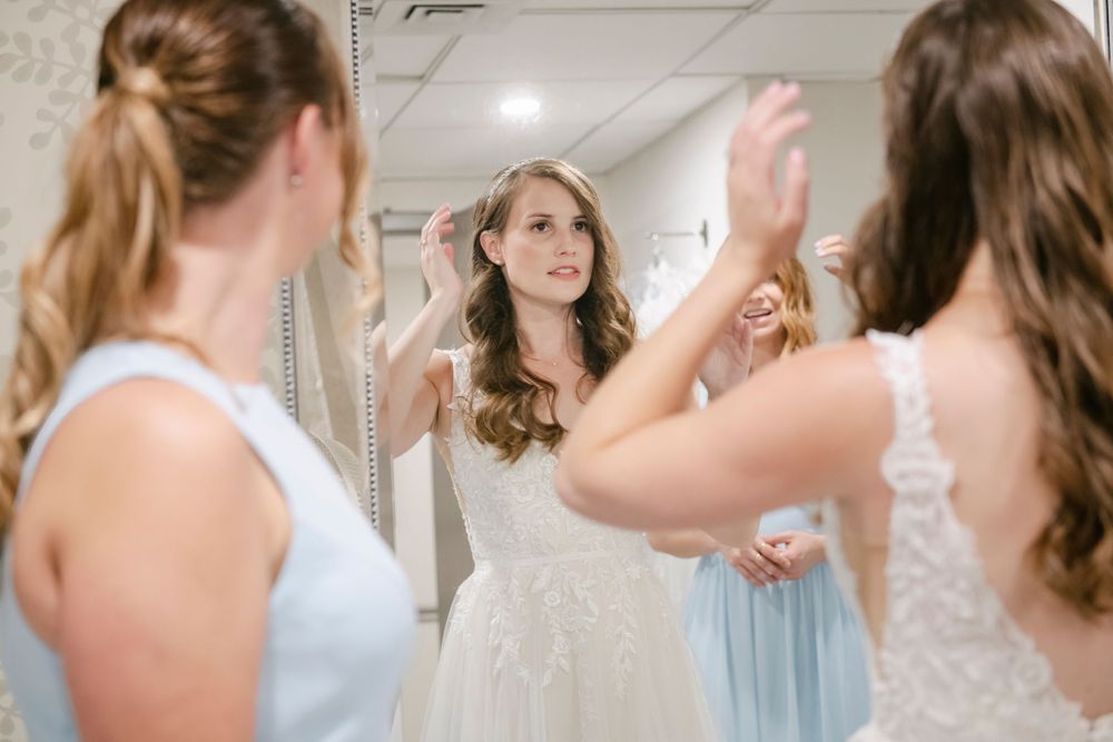 A bride in a white wedding gown looks at her reflection while getting ready with bridesmaids in light blue dresses at Curtis Arboretum