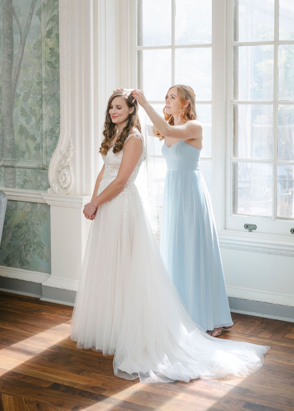 Bridesmaid in light blue dress helps bride with final touches in an elegant room with sunlight streaming through large windows.