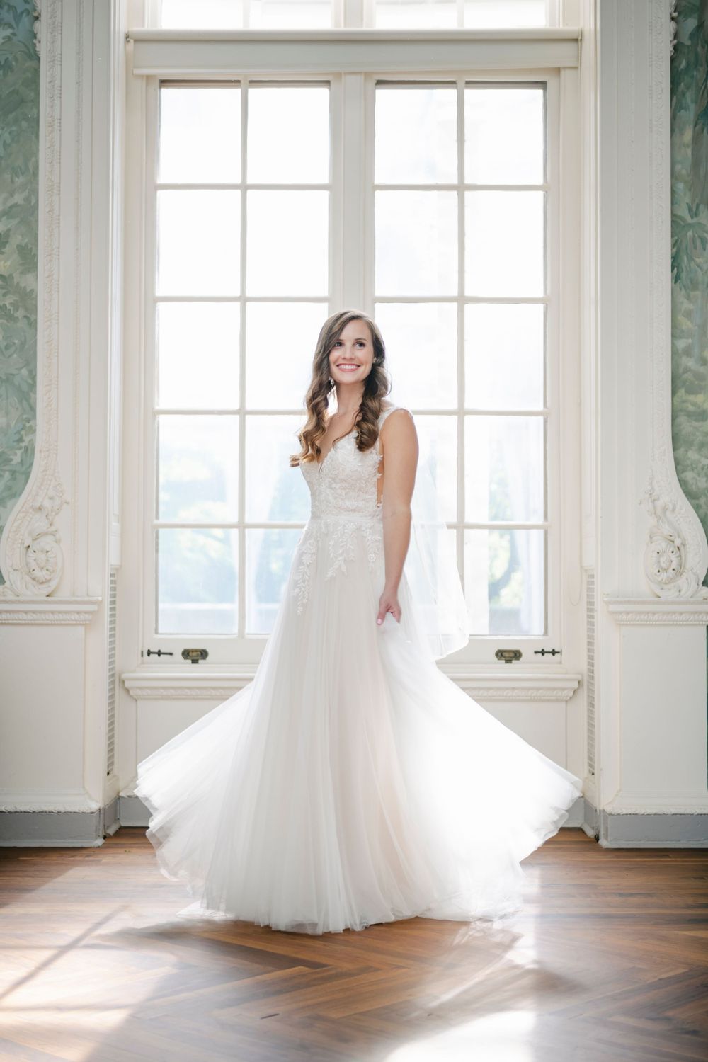 A flowing white wedding gown shimmers in natural light streaming through ornate floor-to-ceiling windows in an elegant room.