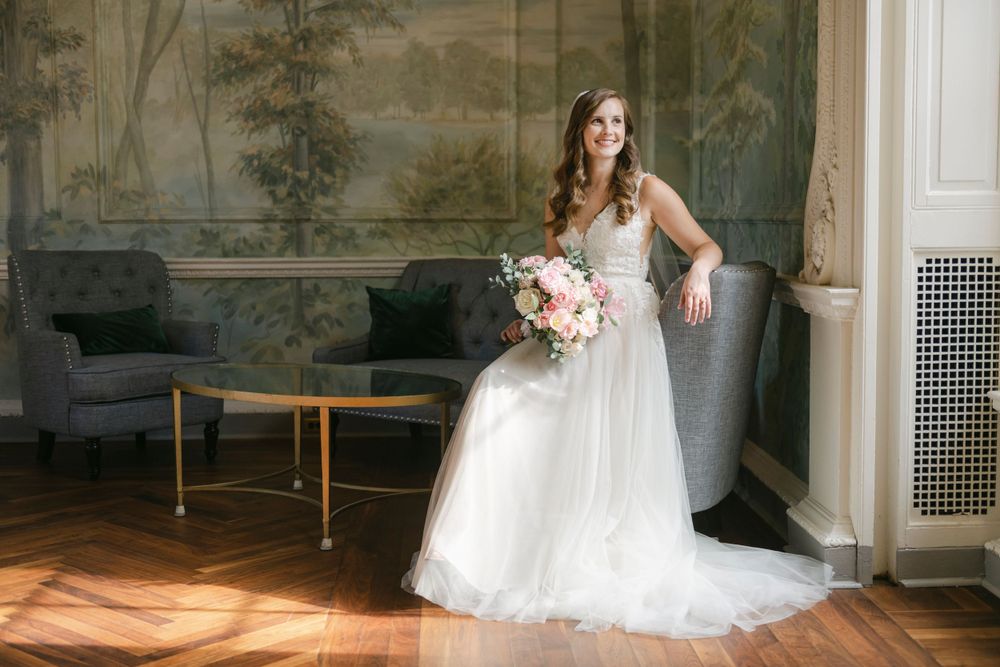 A bride in a flowing white lace gown holds a pink bouquet while posing elegantly in an ornate room.