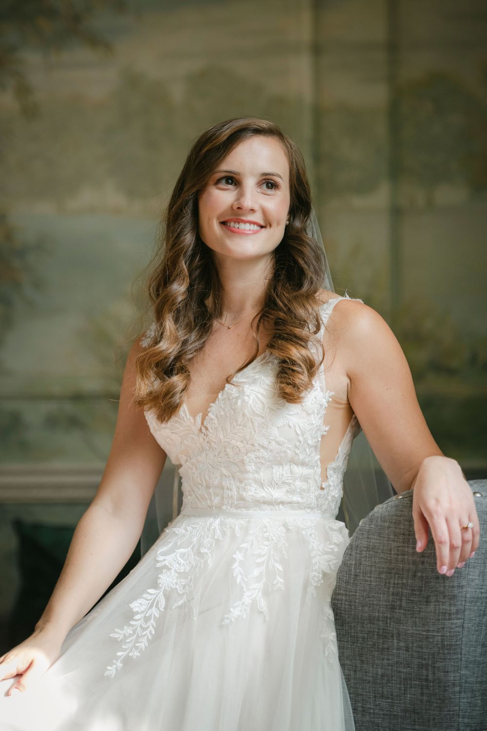 Bride in white lace wedding dress with flowing tulle skirt smiles while sitting on grey chair.