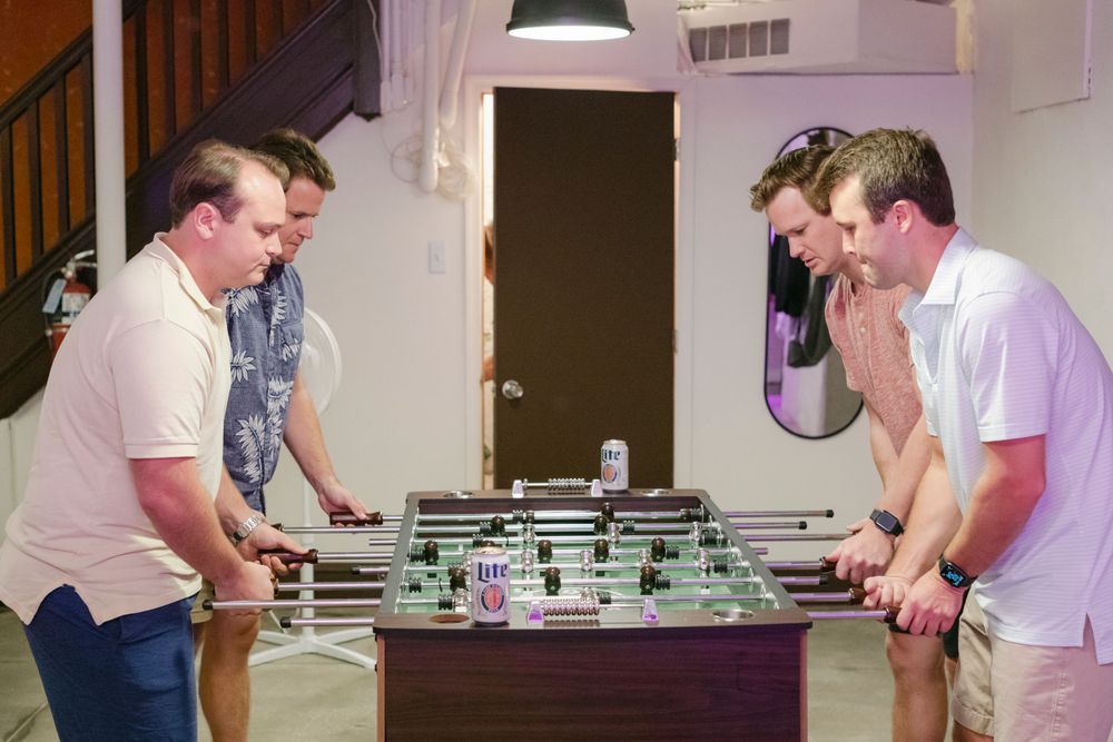 A groom and his groomsmen gather around a foosball table engaged in an intense game of table soccer in an indoor setting.