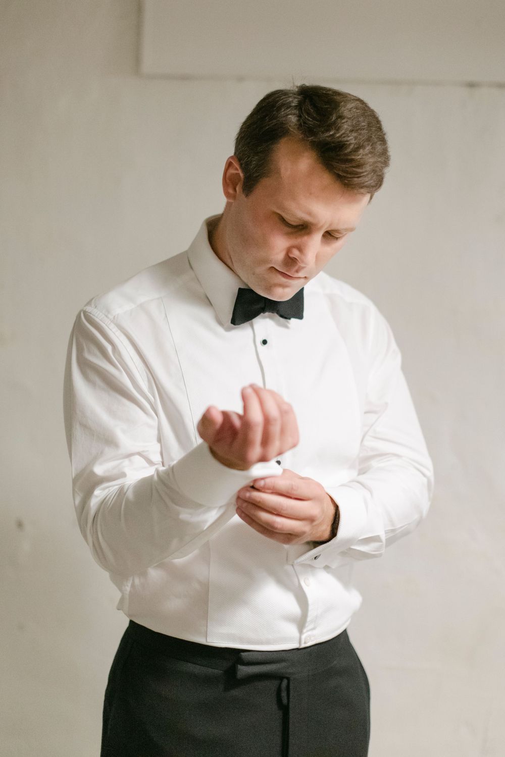 A groom in formal attire with a white dress shirt and black bow tie adjusting his cuff links.