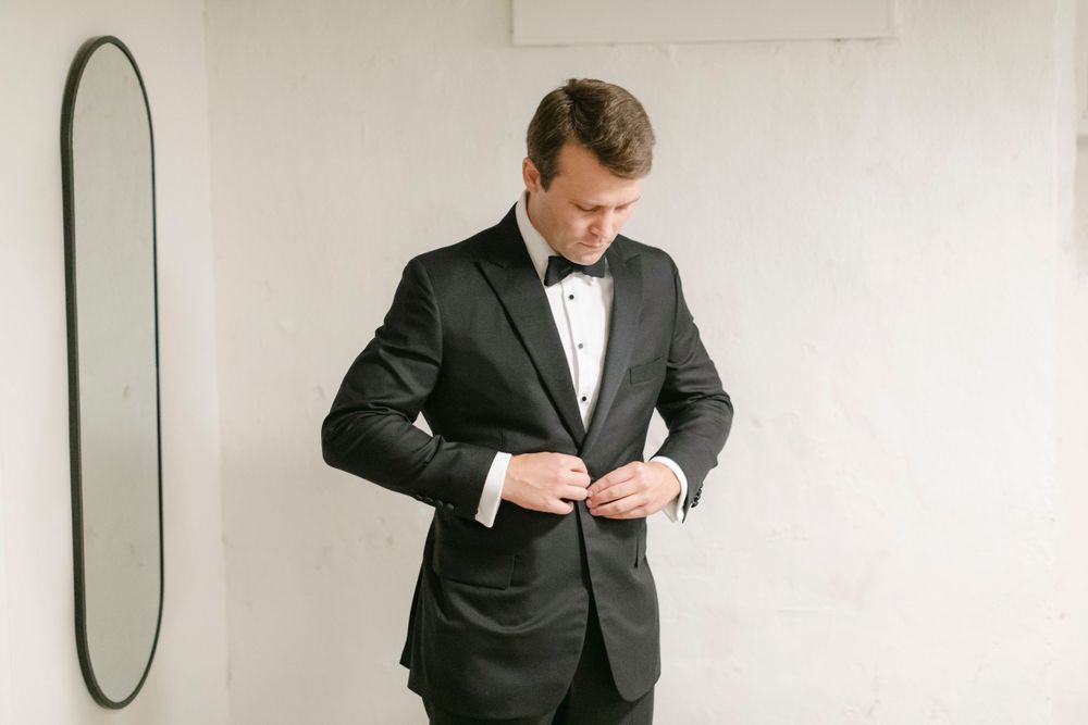 A groom adjusts his classic black tuxedo in a minimalist white room at Curtis Arboretum
