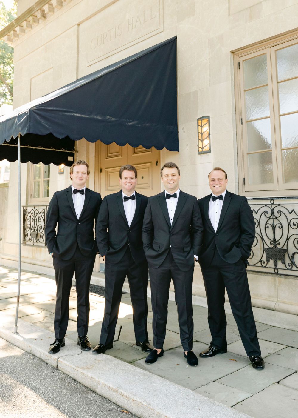 A groom and his groomsmen in formal black tuxedos and bow ties stand together on an elegant stone entrance porch with a dark awning.
