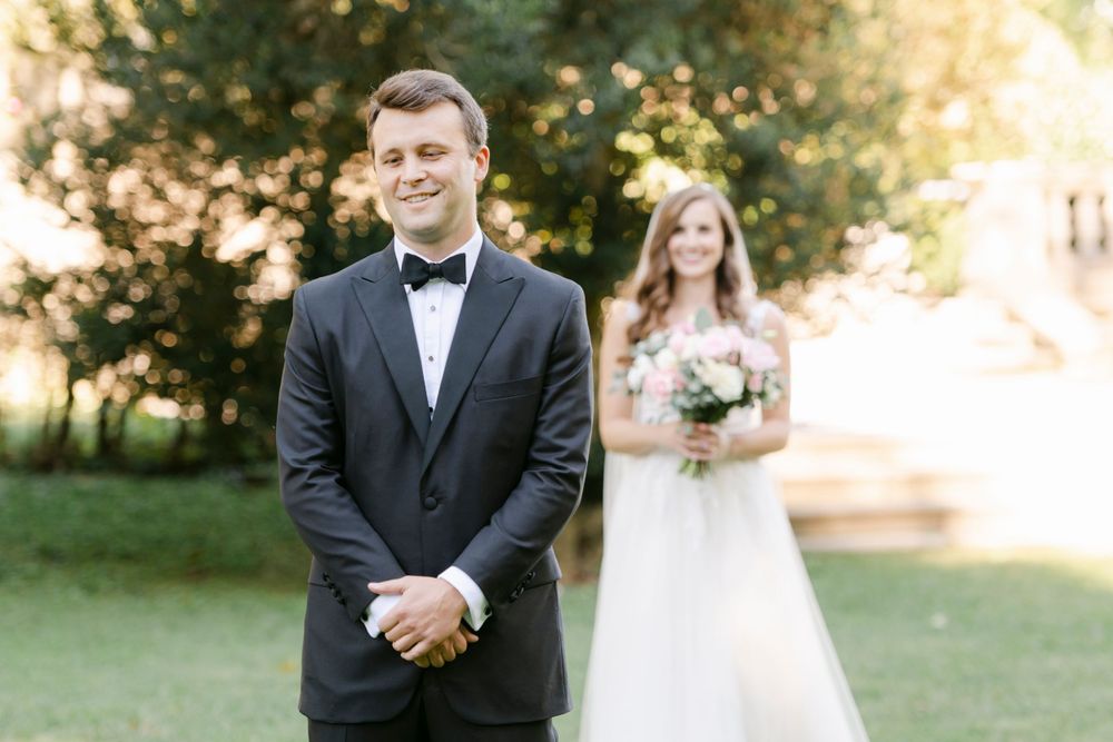 Wedding photo of the first look moment between the groom in a black tuxedo and a bride in white dress with bouquet in sunny garden setting.