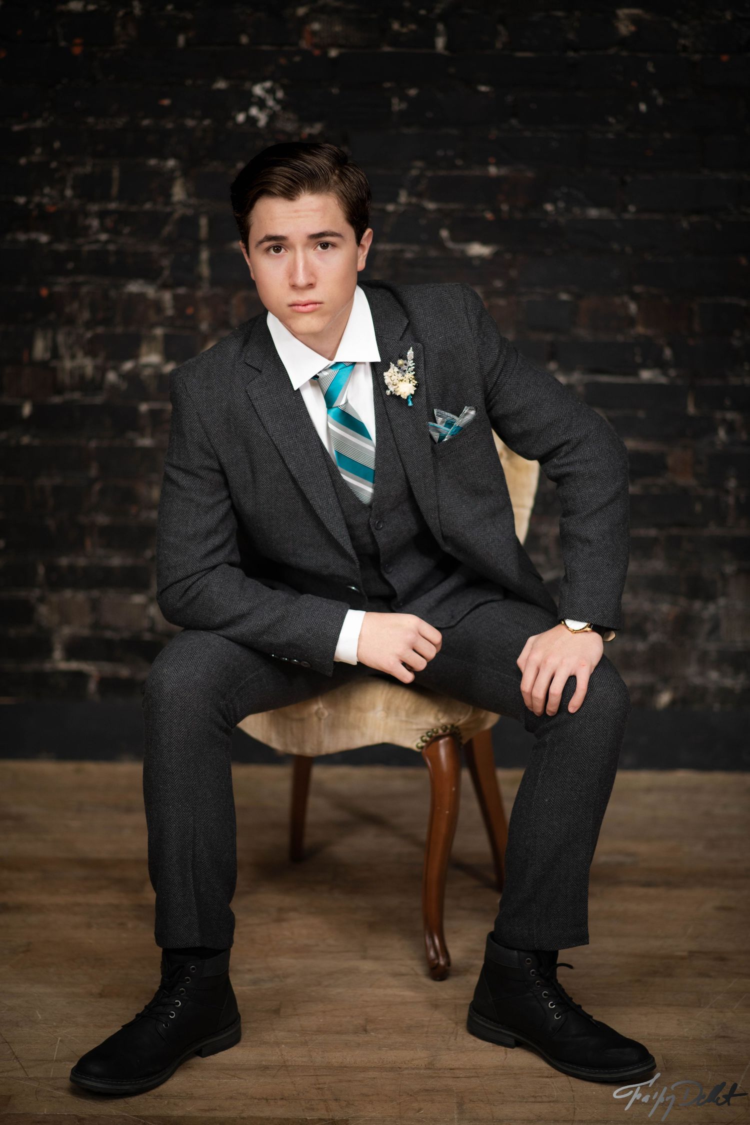 A person in a dark suit with turquoise tie sits on wooden furniture against a rustic brick wall backdrop.