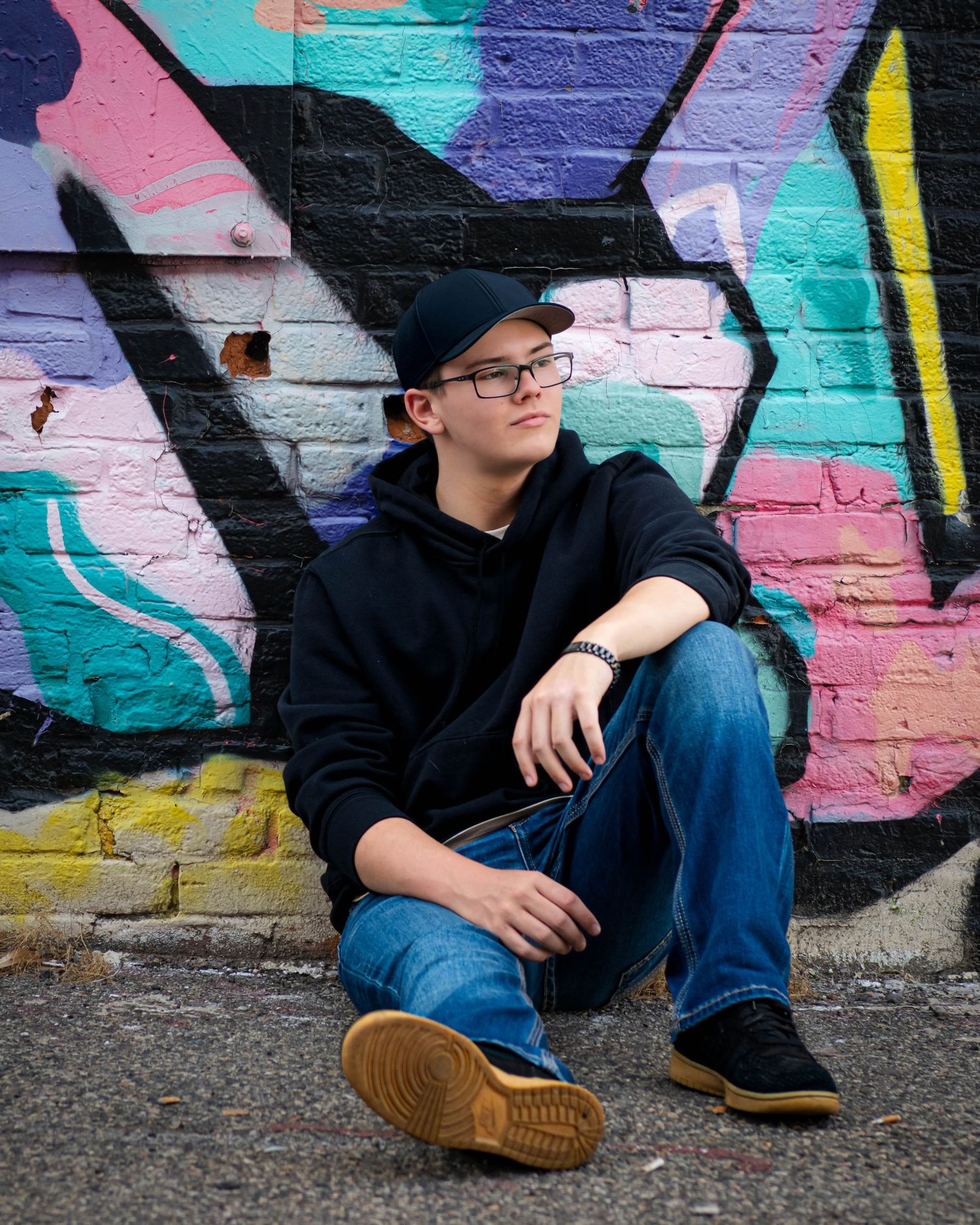 A person in dark clothing sits against a colorful graffiti wall wearing brown shoes and jeans.