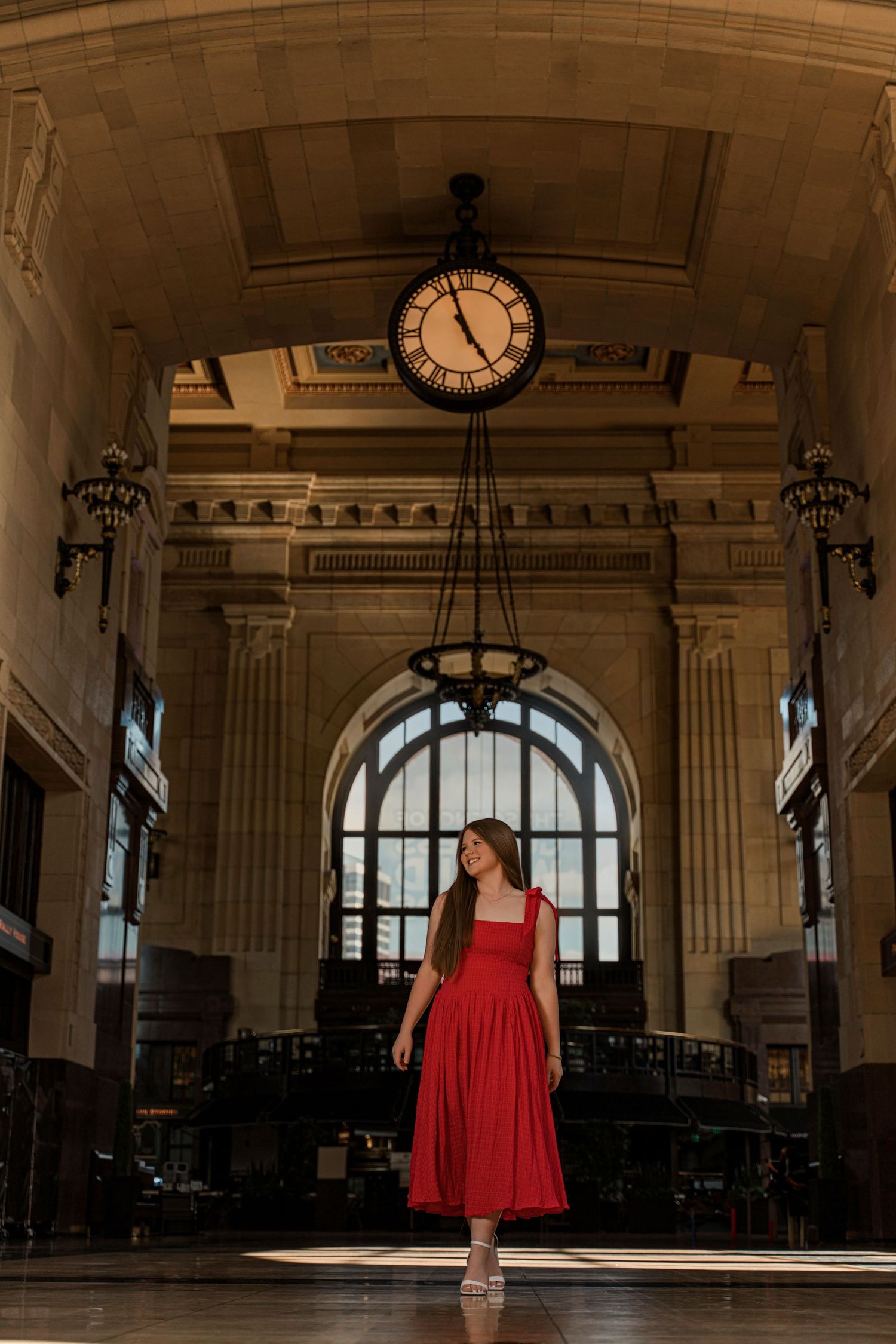 A woman in a red dress stands in an elegant train station hall with a vintage clock and arched architecture.