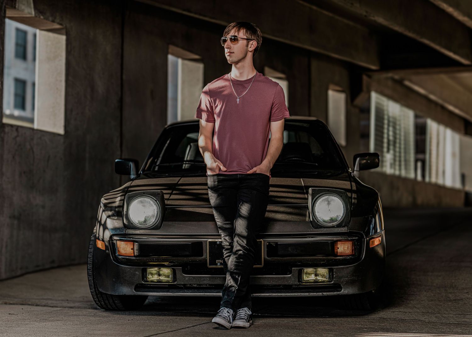 A person stands next to a vintage black sports car in an industrial parking garage setting.