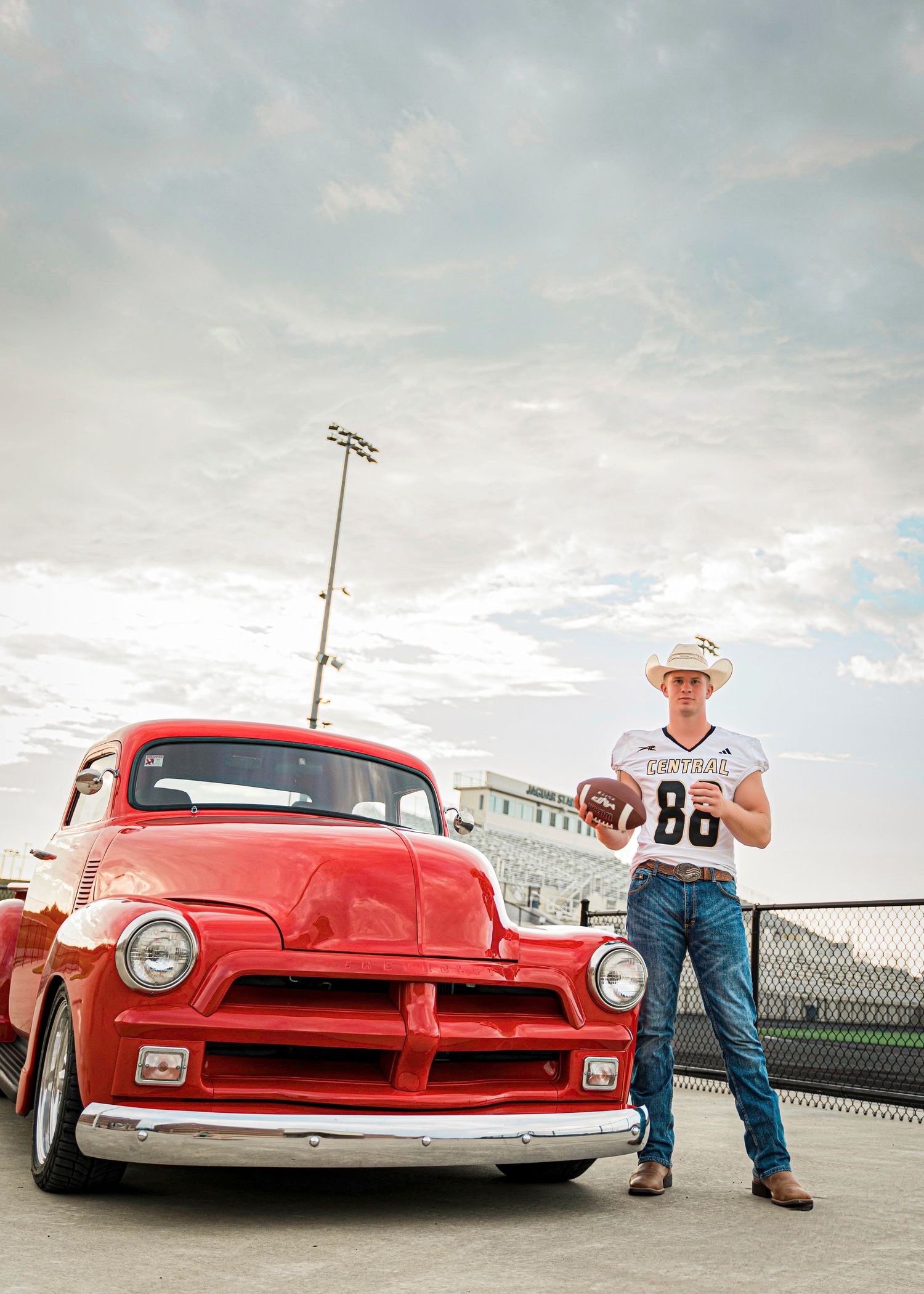 A bright red custom hot rod car with lowered suspension sits in front of stadium lights at dusk.