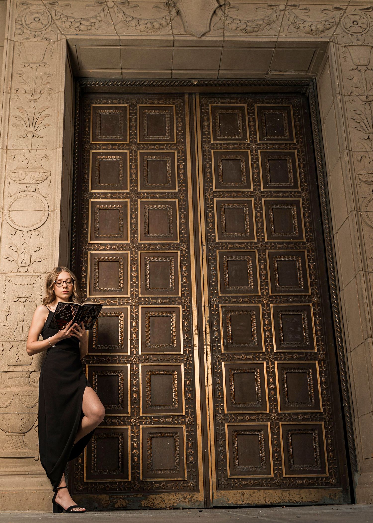 A black dress contrasts against ornate bronze doors with geometric patterns in an elegant architectural setting.