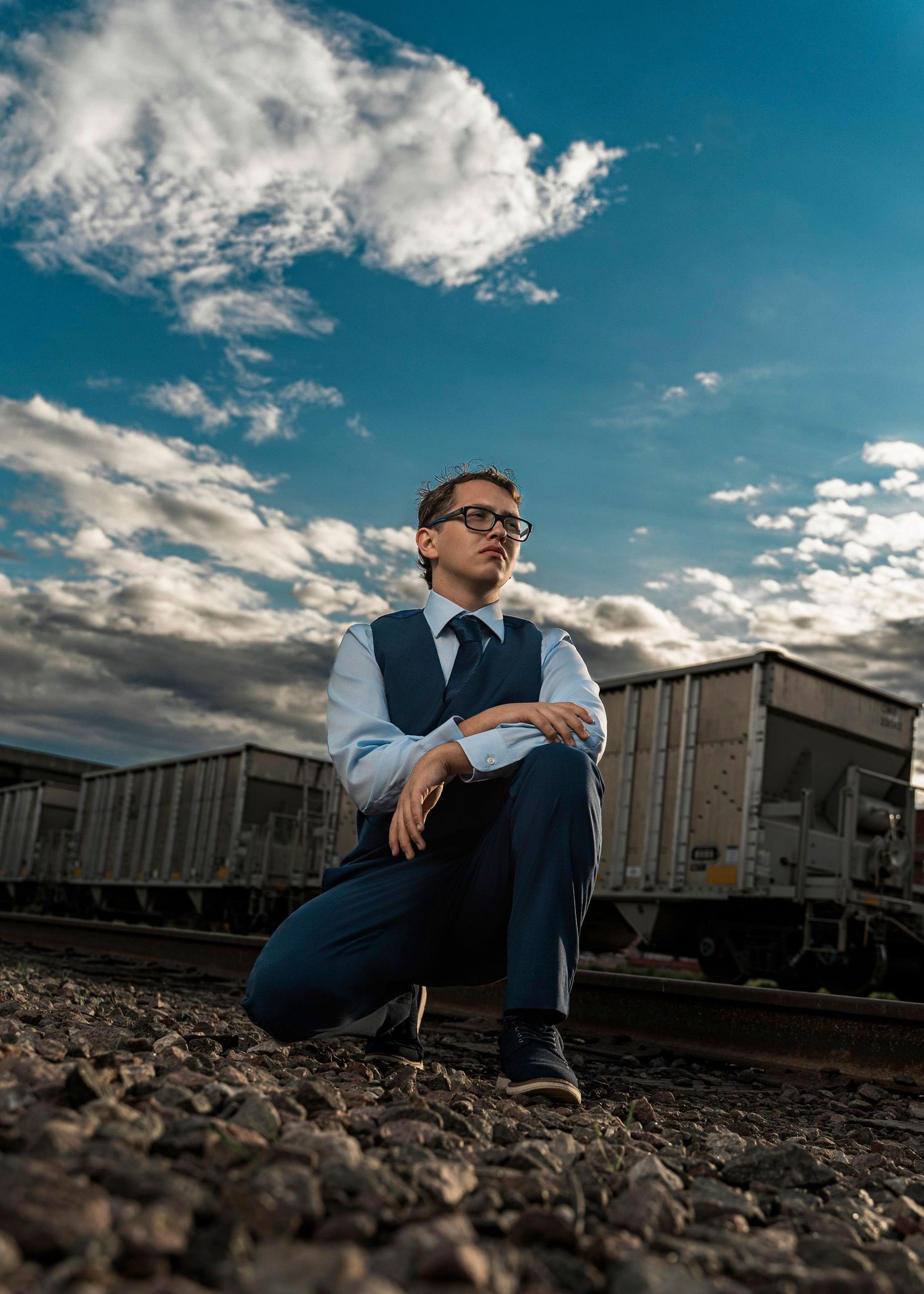 A person in business attire sits thoughtfully on train tracks against a dramatic cloudy sky at sunset.