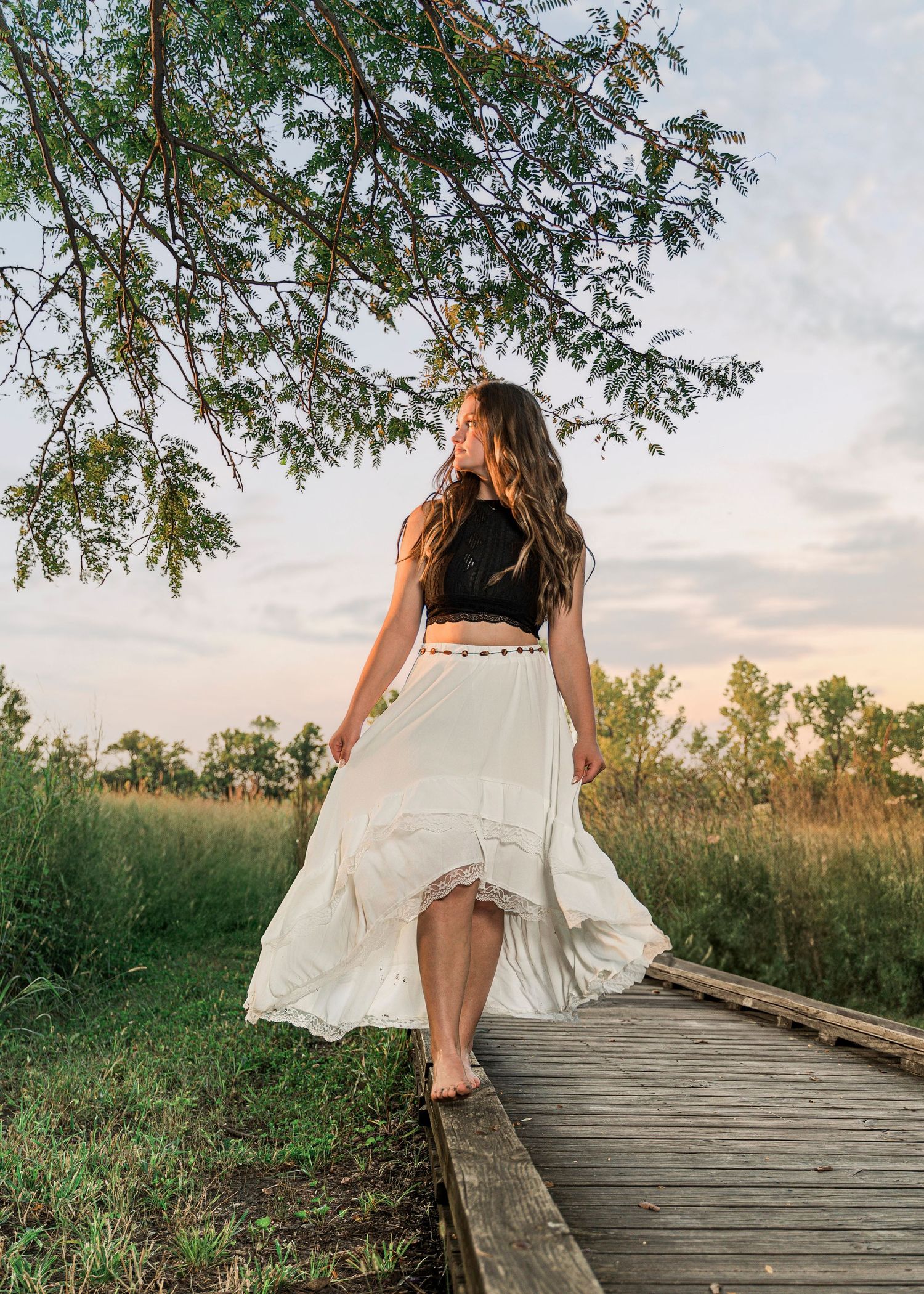 A flowing white skirt billows in the breeze as someone walks along a wooden boardwalk at sunset beneath tree branches.