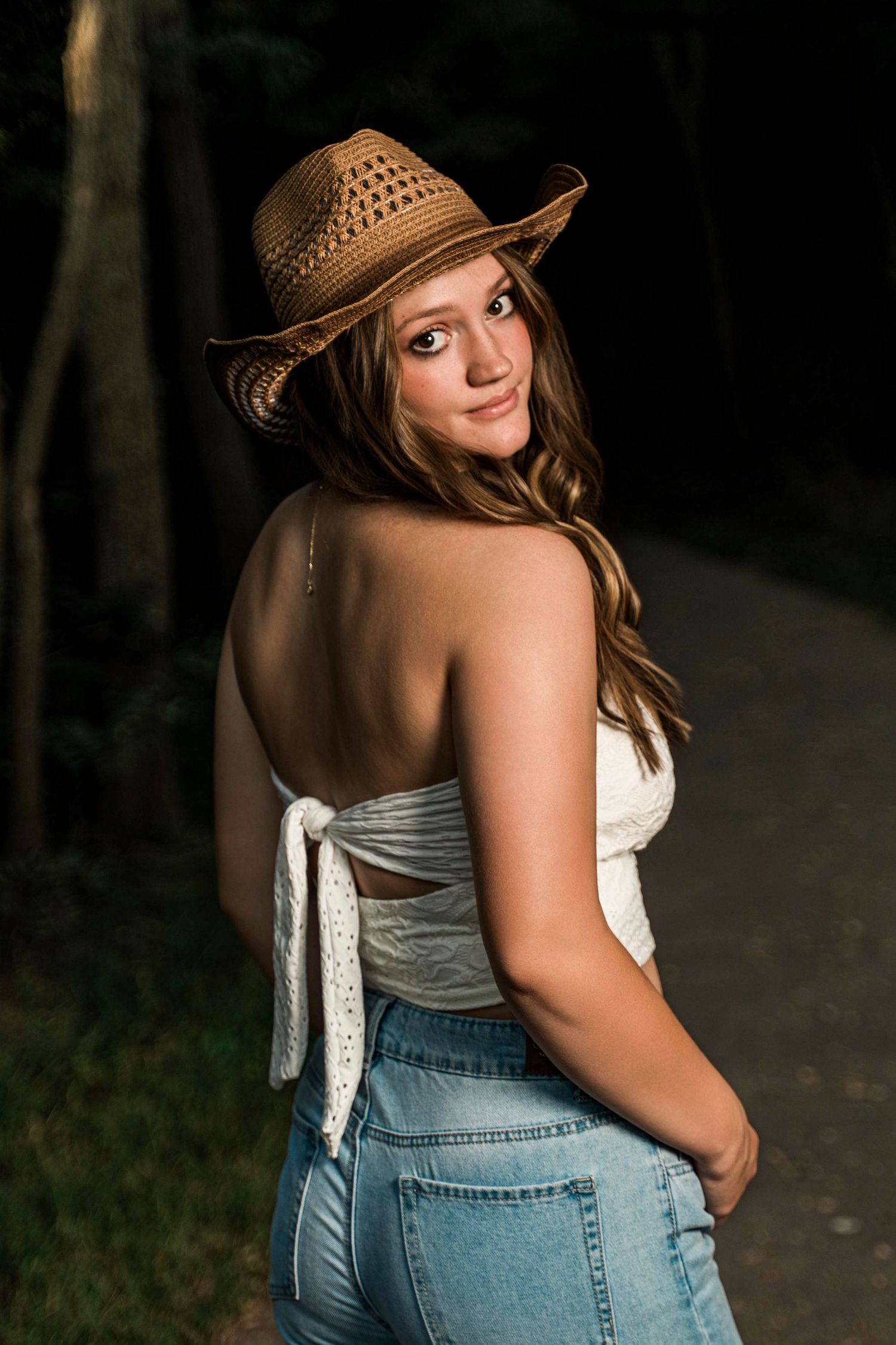 A woman in a straw hat and denim overalls poses outdoors against a dark background.