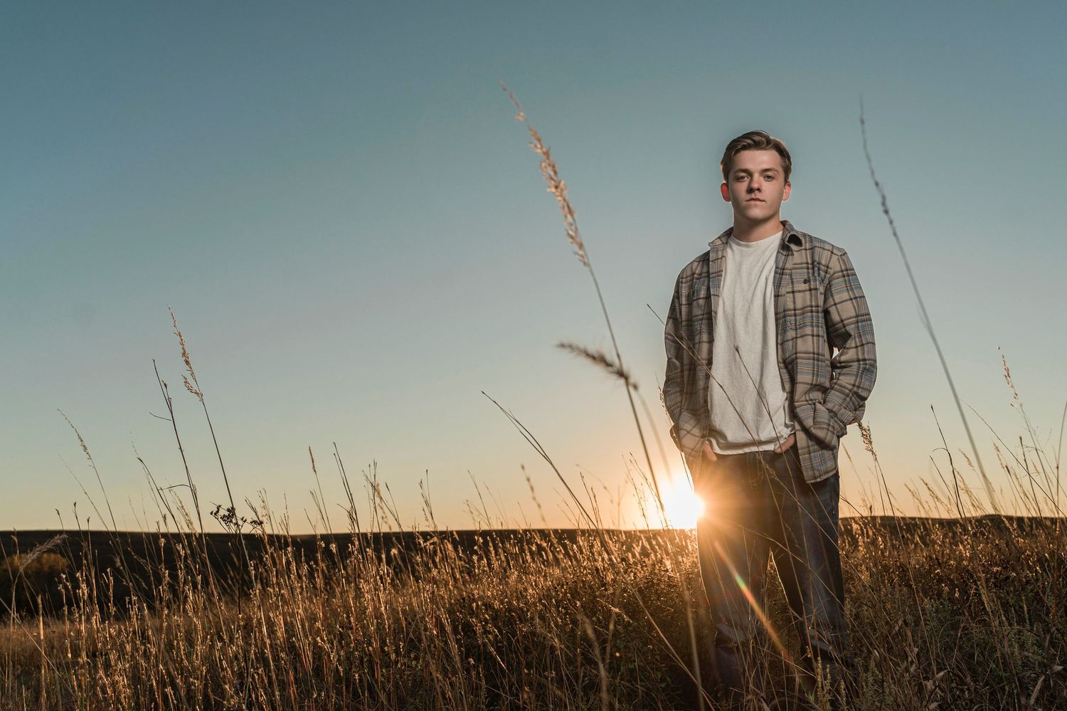 A person stands in a golden wheat field at sunset wearing a plaid jacket against the warm evening sky.