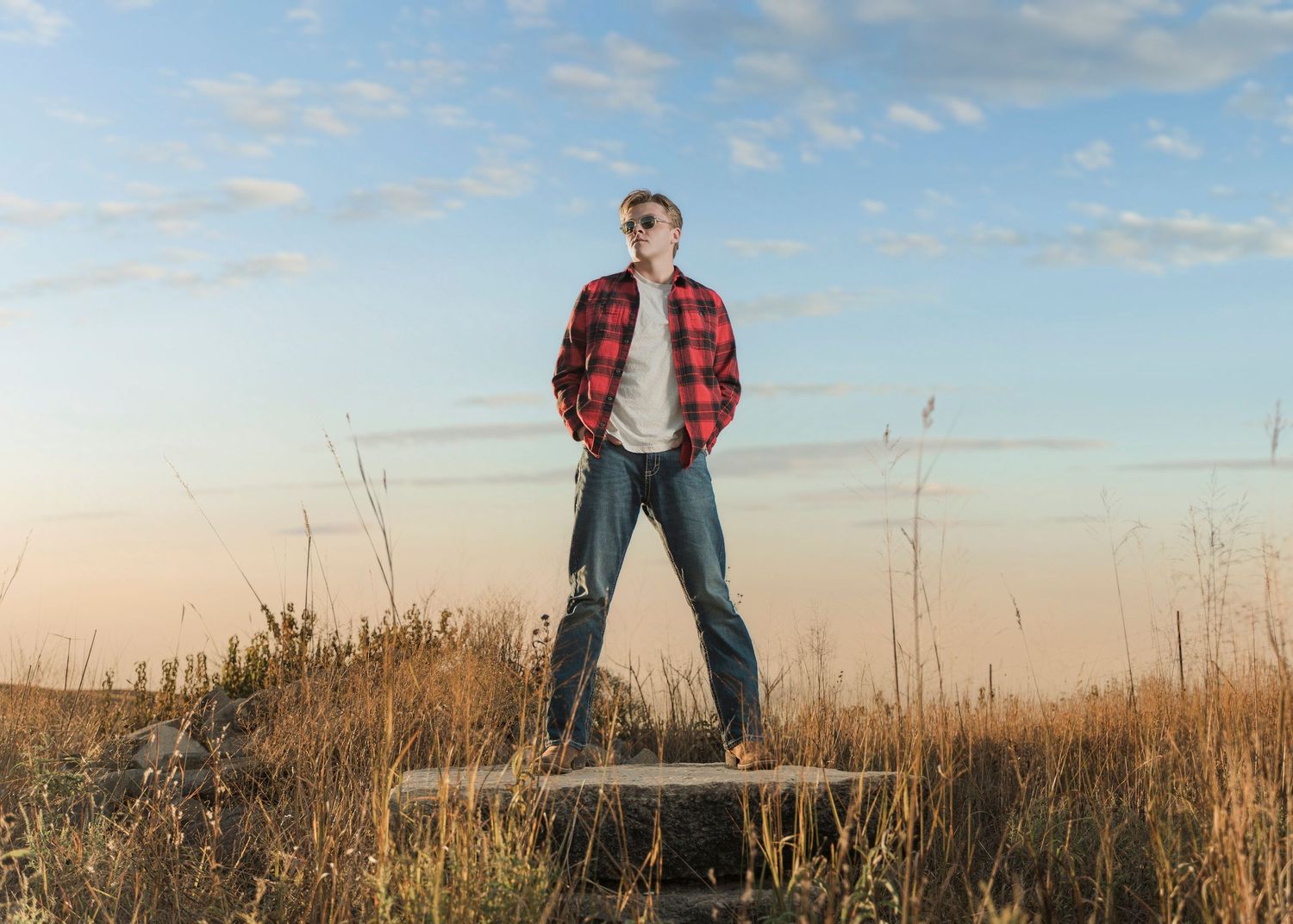 A person in a red plaid jacket and jeans stands in a golden field of tall grass at sunset.