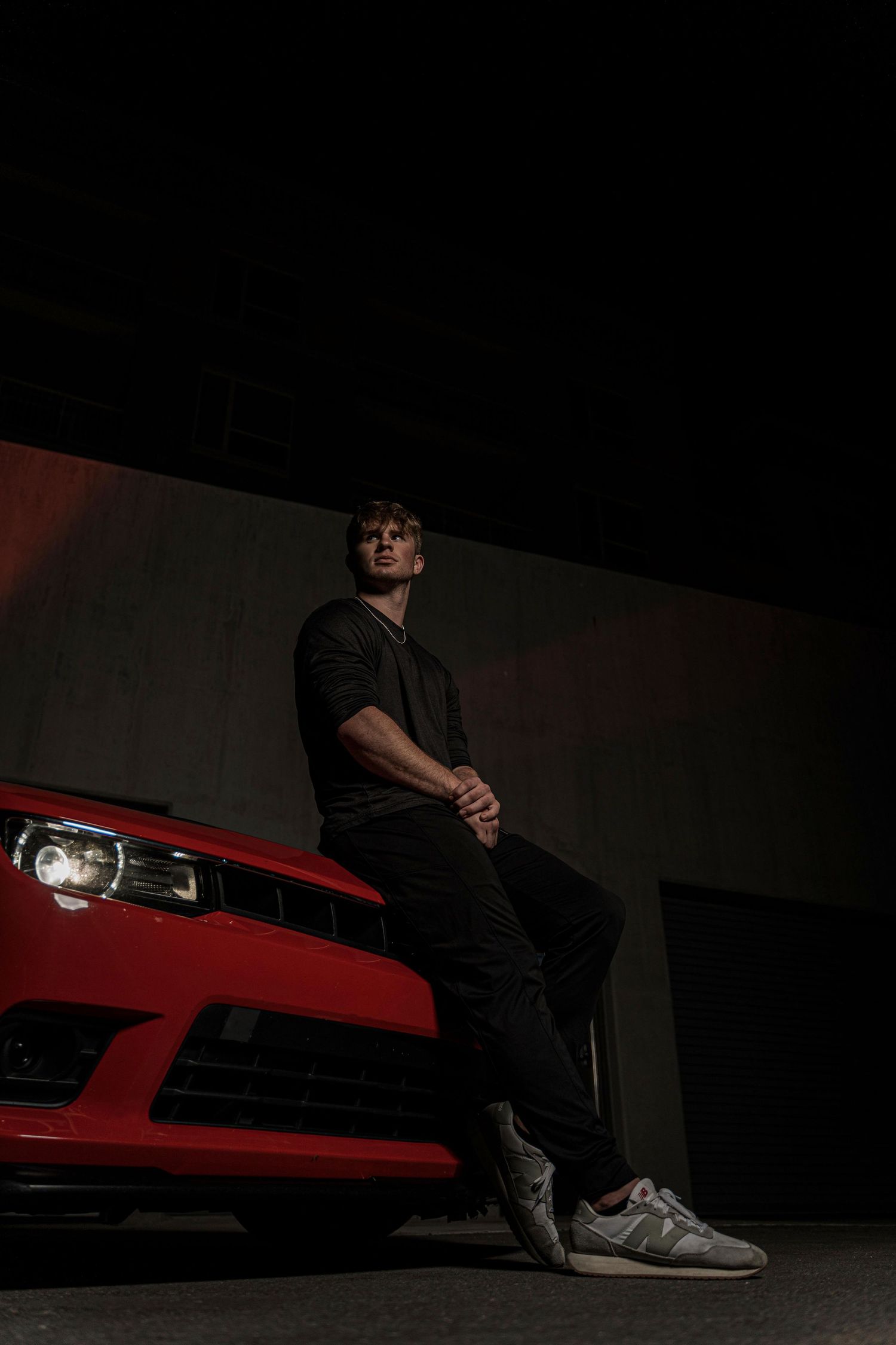 Moody nighttime portrait of someone in black clothing leaning against a red sports car in dramatic low lighting.