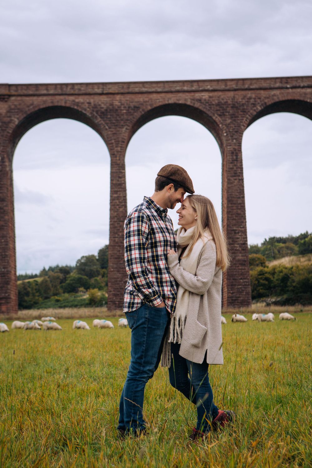 A couple shares an intimate moment beneath a brick viaduct with arches while standing in a grassy meadow.