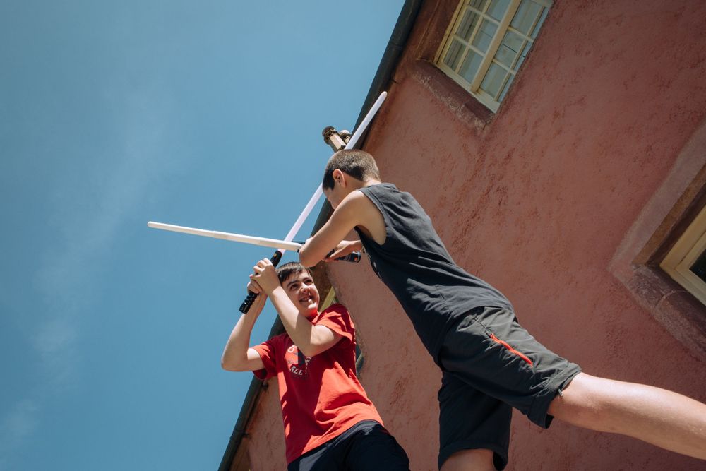 Two people pose dramatically against a blue sky and building wall while practicing stage sword fighting.