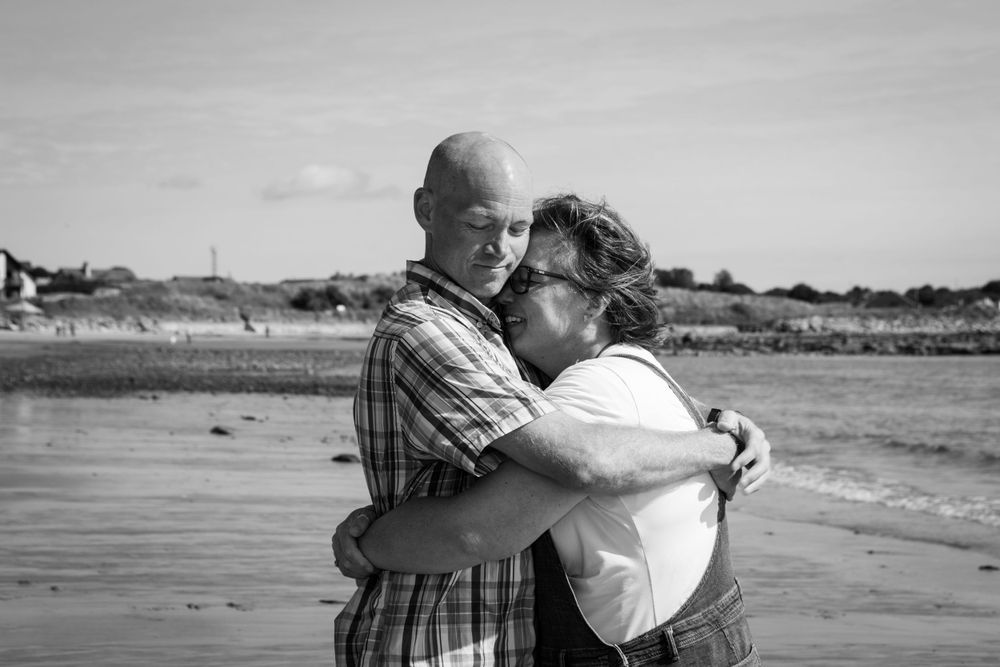 A loving couple embraces on a beach in black and white, with waves and sandy shore in the background.