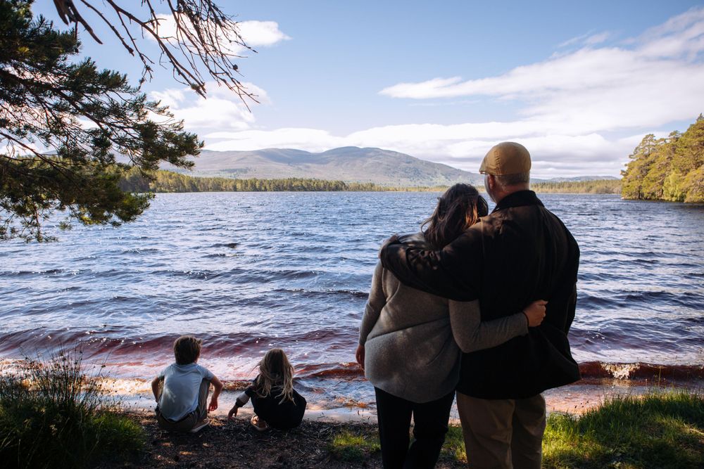 People stand together enjoying the view of a serene lake surrounded by mountains on a sunny day.