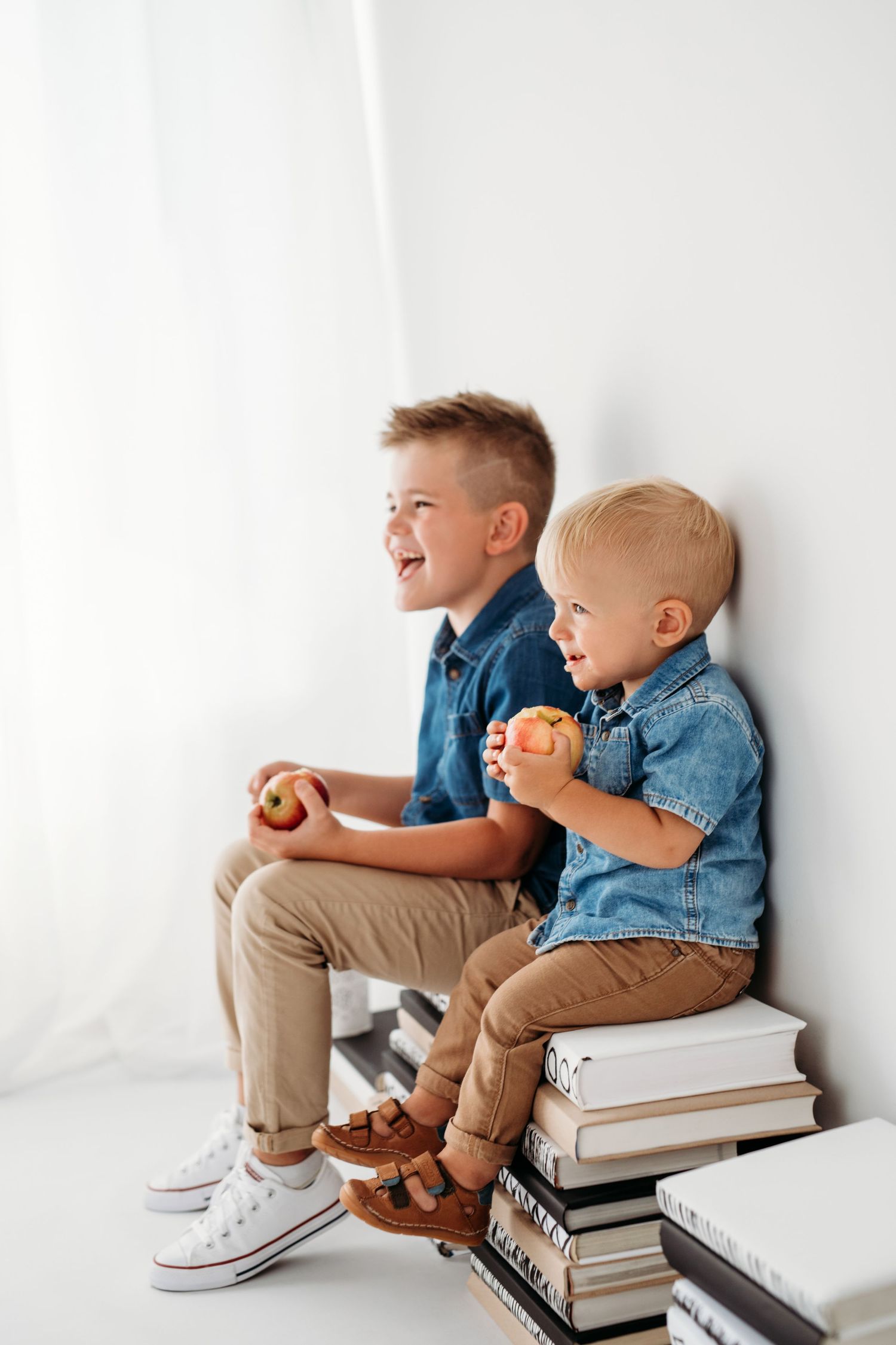 Two children sitting on stack of books wearing denim shirts and holding apples.
