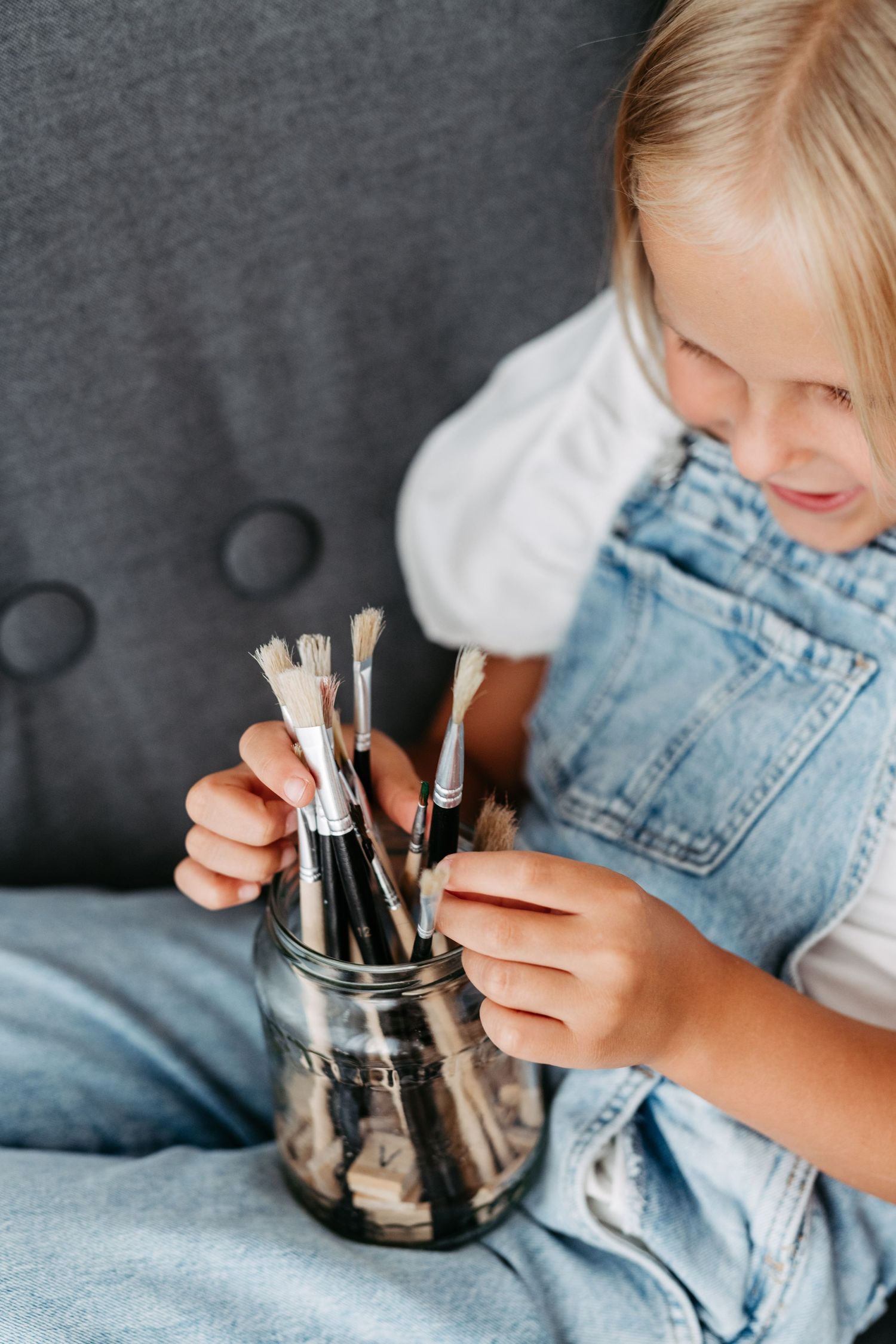 Close-up of small hands holding paint brushes from glass jar on grey chair.