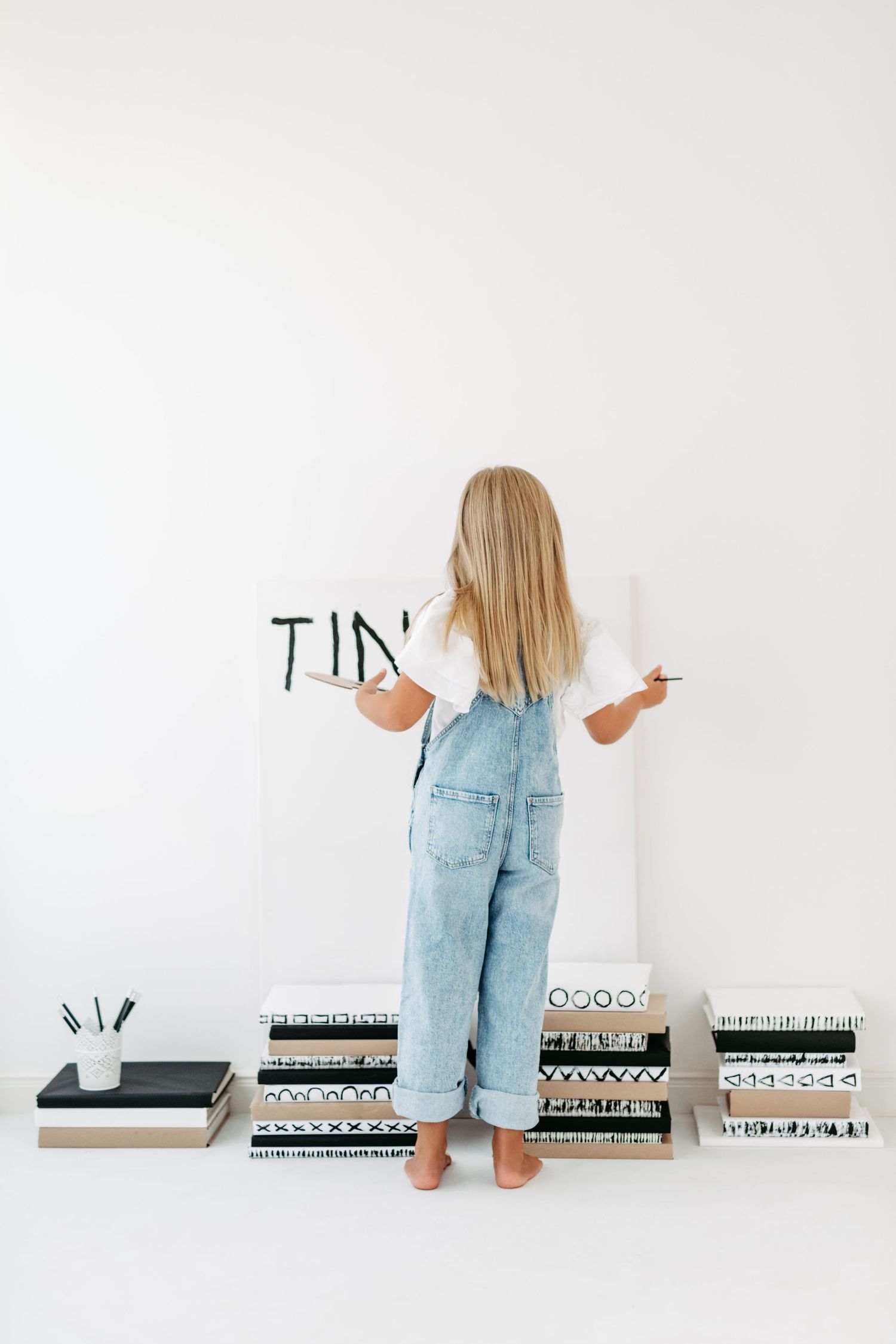 Child in denim overalls writing on wall with art supplies displayed below.