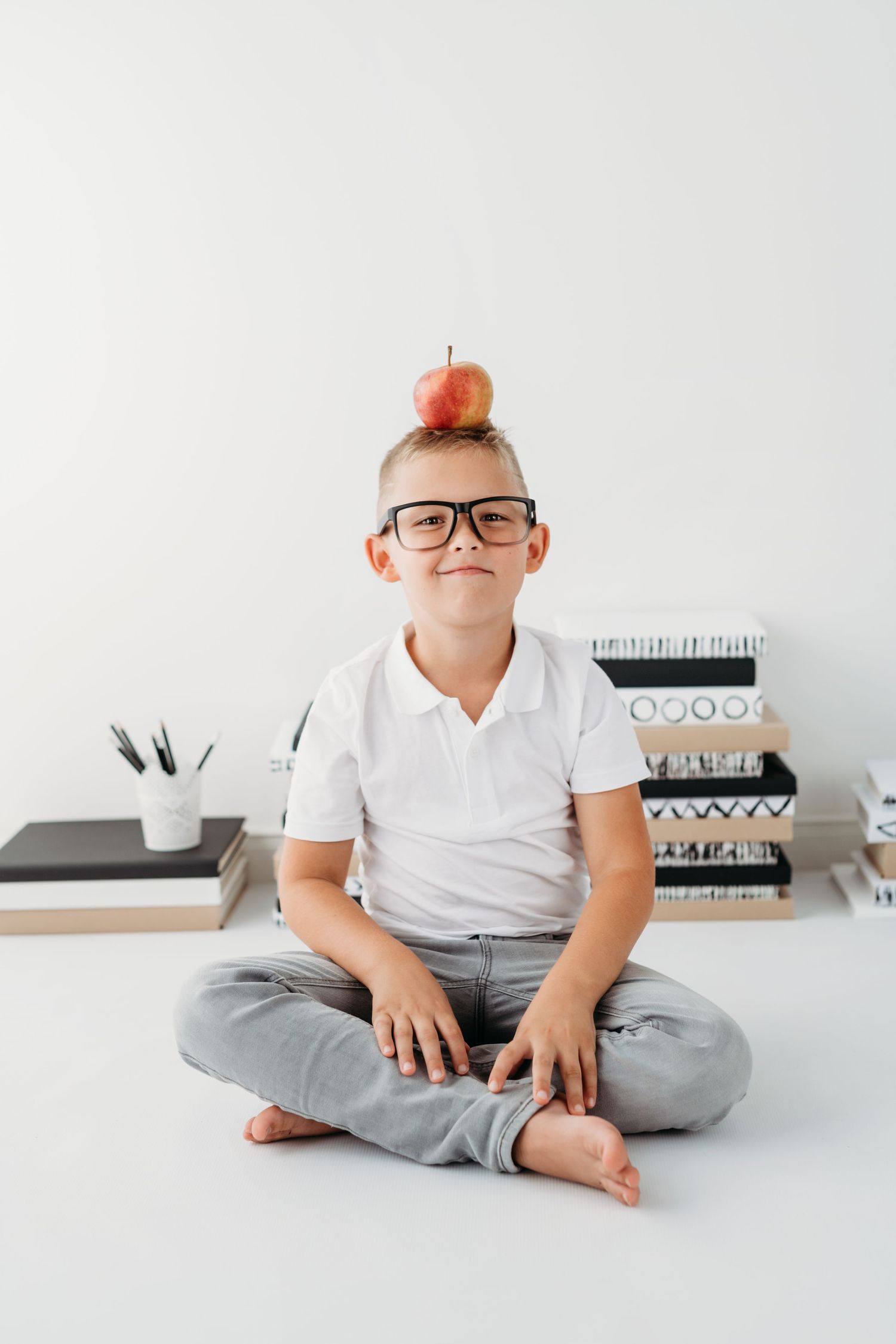 Child in glasses sitting cross-legged with red apple balanced on head against white background.