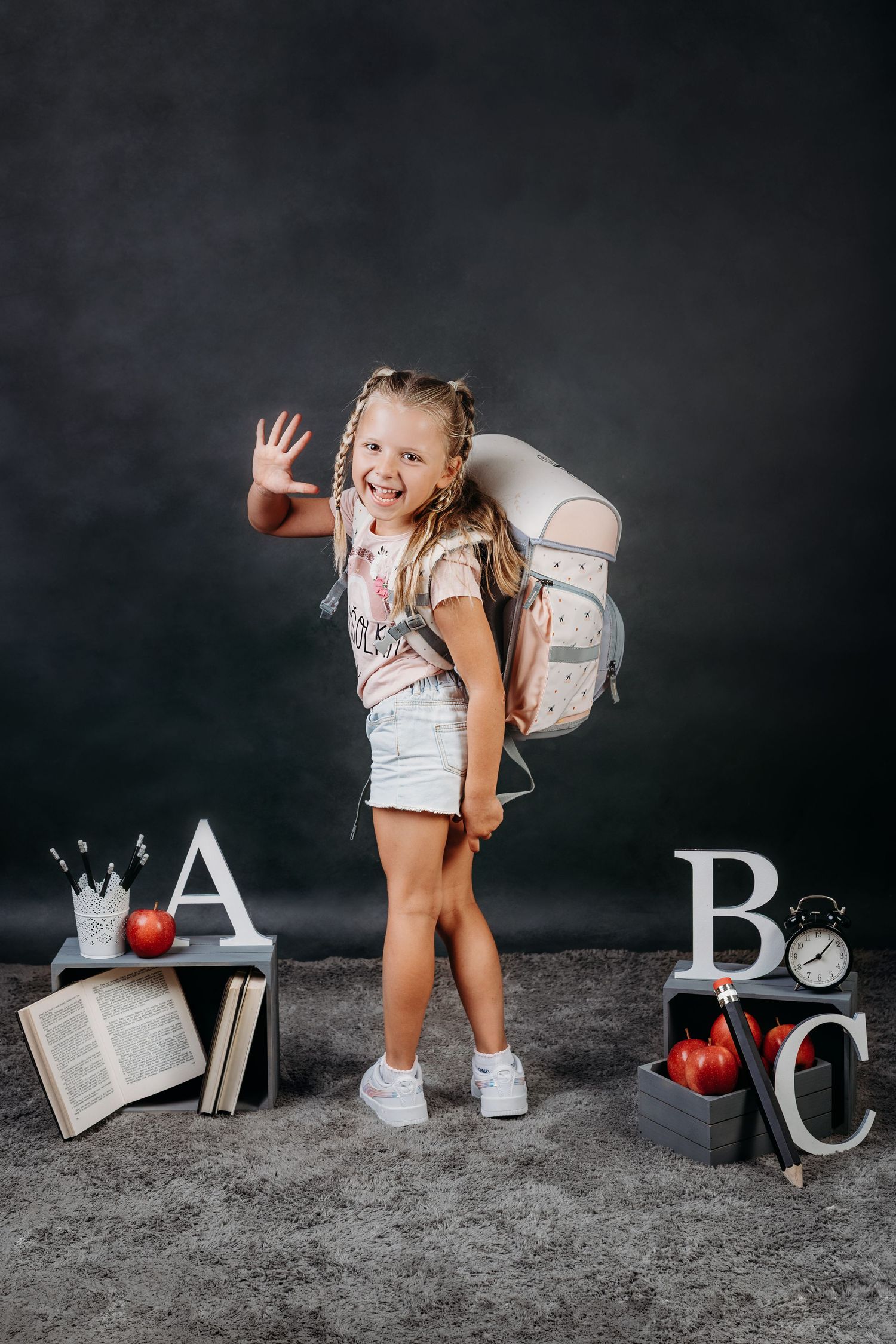A cheerful young girl with braids waves while wearing a backpack, surrounded by ABC letters, books, and apples on a dark background.