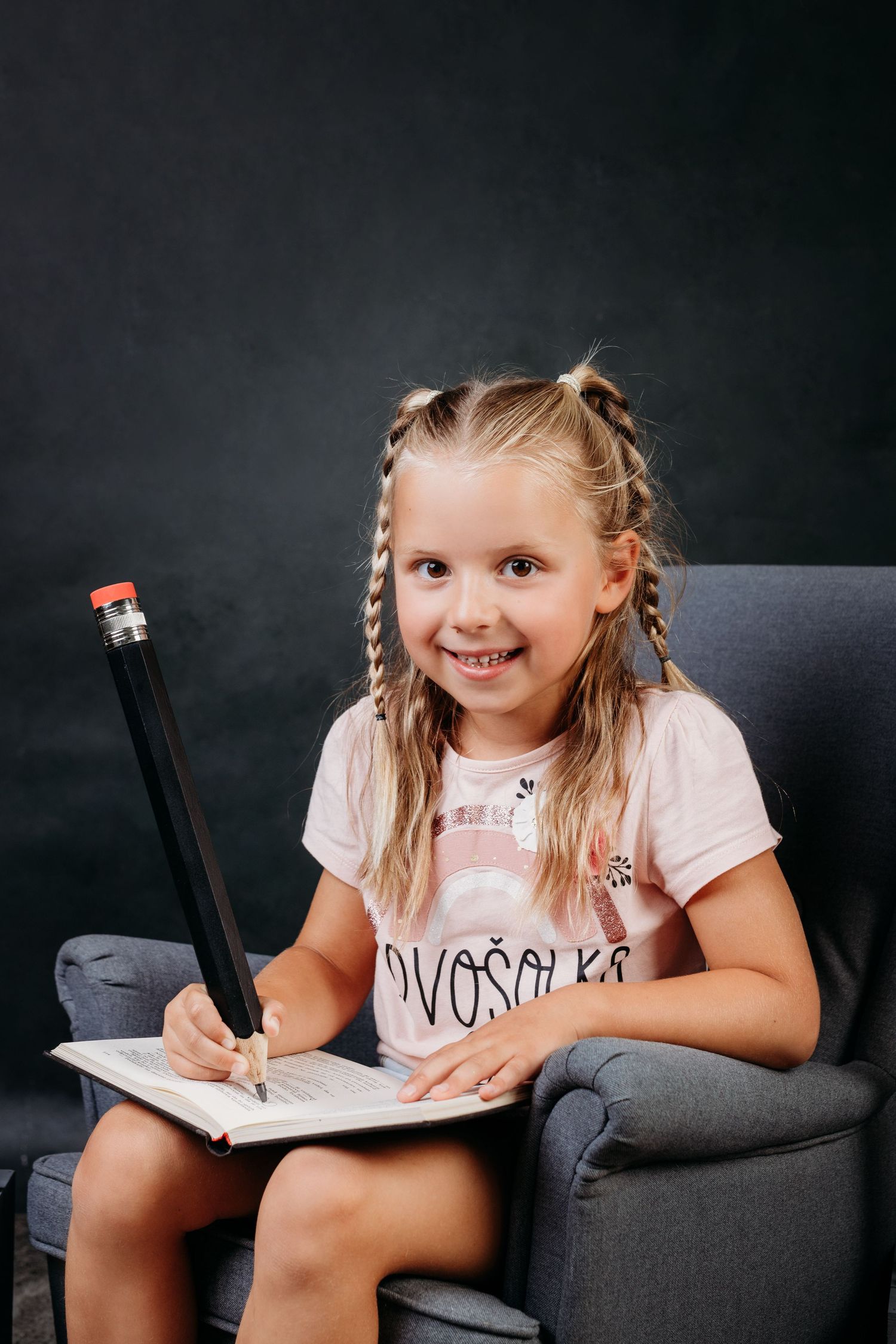 Smiling young girl sitting in chair holding oversized pencil and writing in notebook.