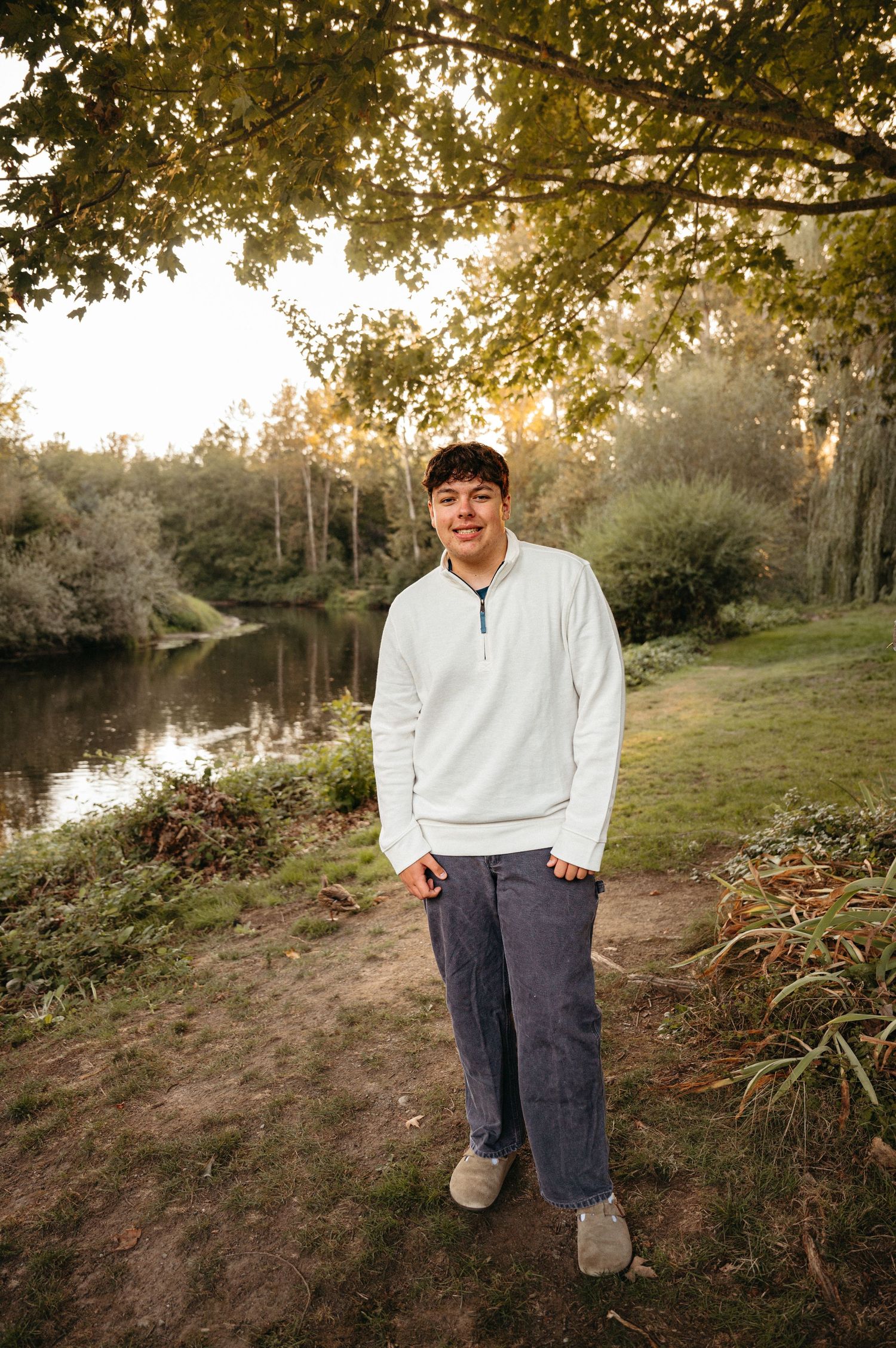 Person in white sweater standing by serene pond at sunset with autumn trees overhead.