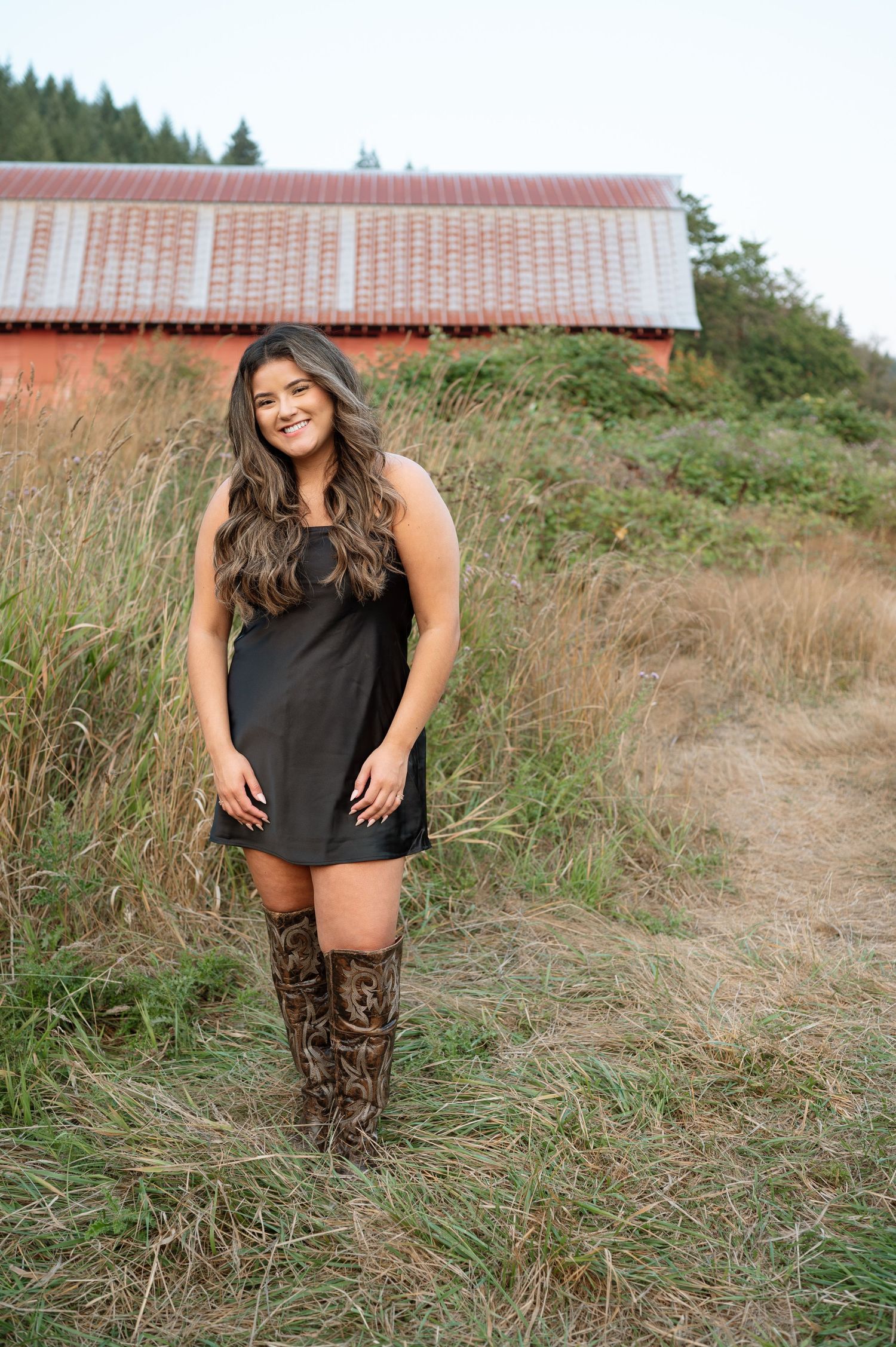Person in black dress and brown boots posing in field with red barn in background.