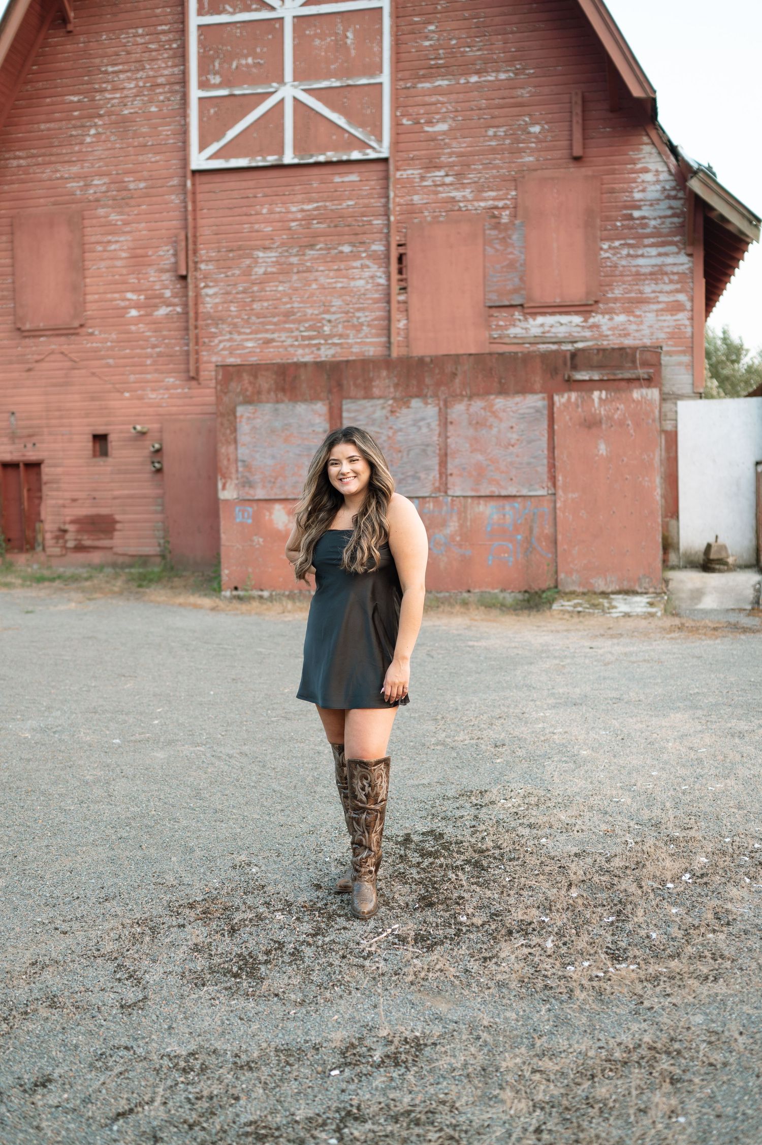 Person in black dress and boots standing in front of rustic red barn.