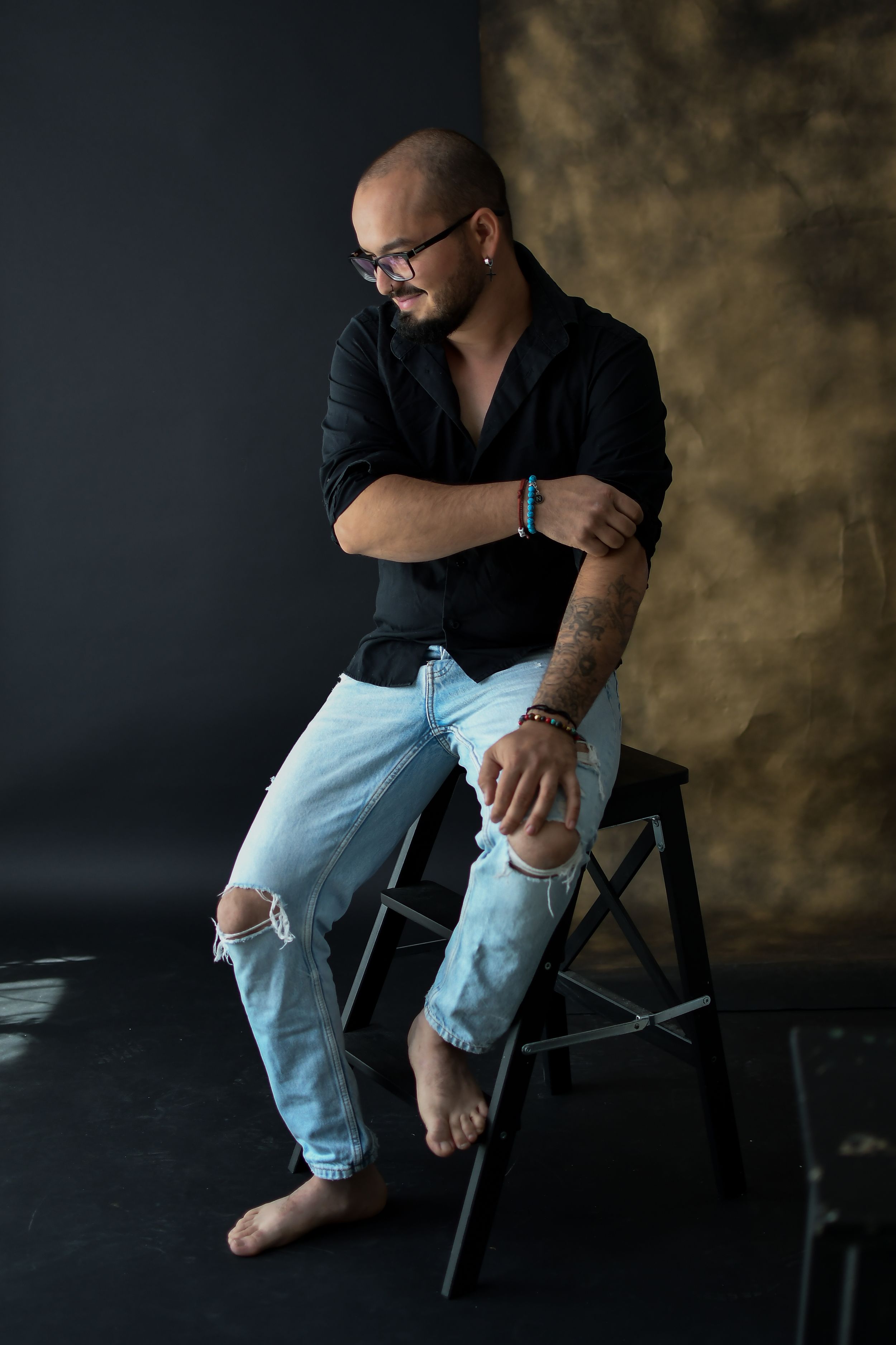 Artistic studio portrait of someone in a black shirt and ripped jeans seated barefoot on a stool against dark background.