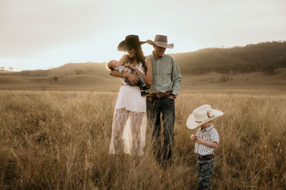 Family in cowboy hats standing in golden grass field at sunset, parents holding newborn baby with young child nearby.